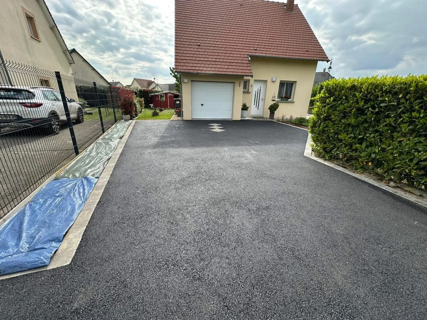 Une maison avec un garage et une allée récemment asphaltée, entourée d'un mur en haie et d'une clôture métallique, avec des voitures stationnées à côté.