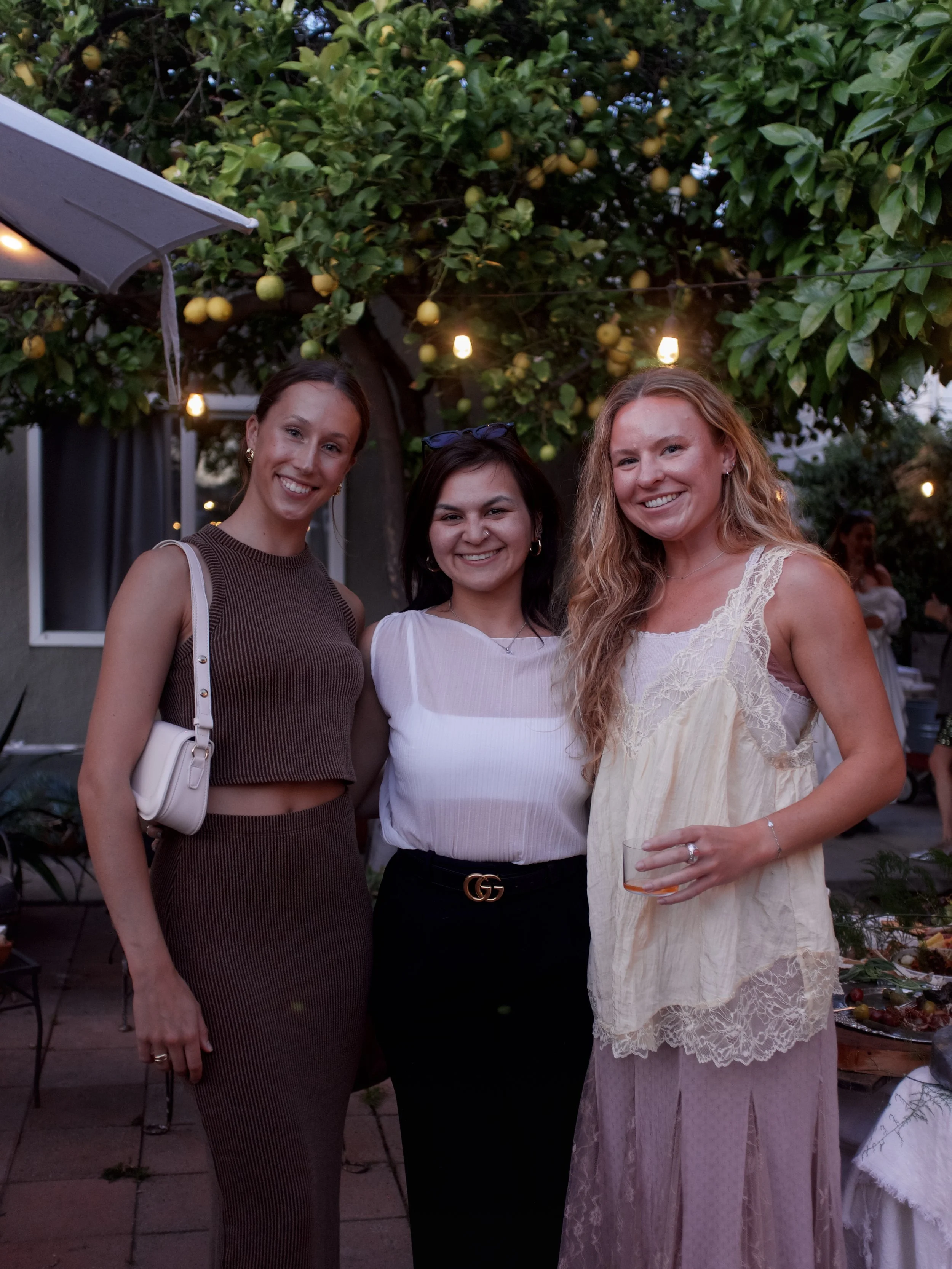 Three women standing outdoors at twilight, smiling at the camera. They are dressed casually, with a lemon tree and string lights in the background.
