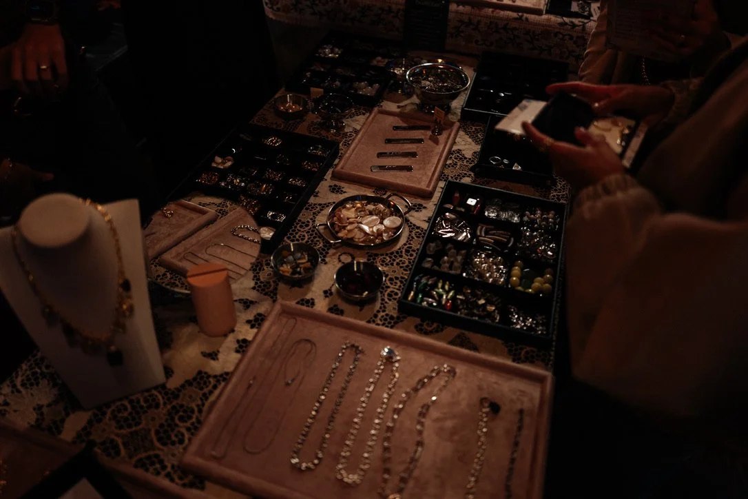 Jewelry display table with necklaces, rings, and other accessories, with people browsing and photographing the jewelry.