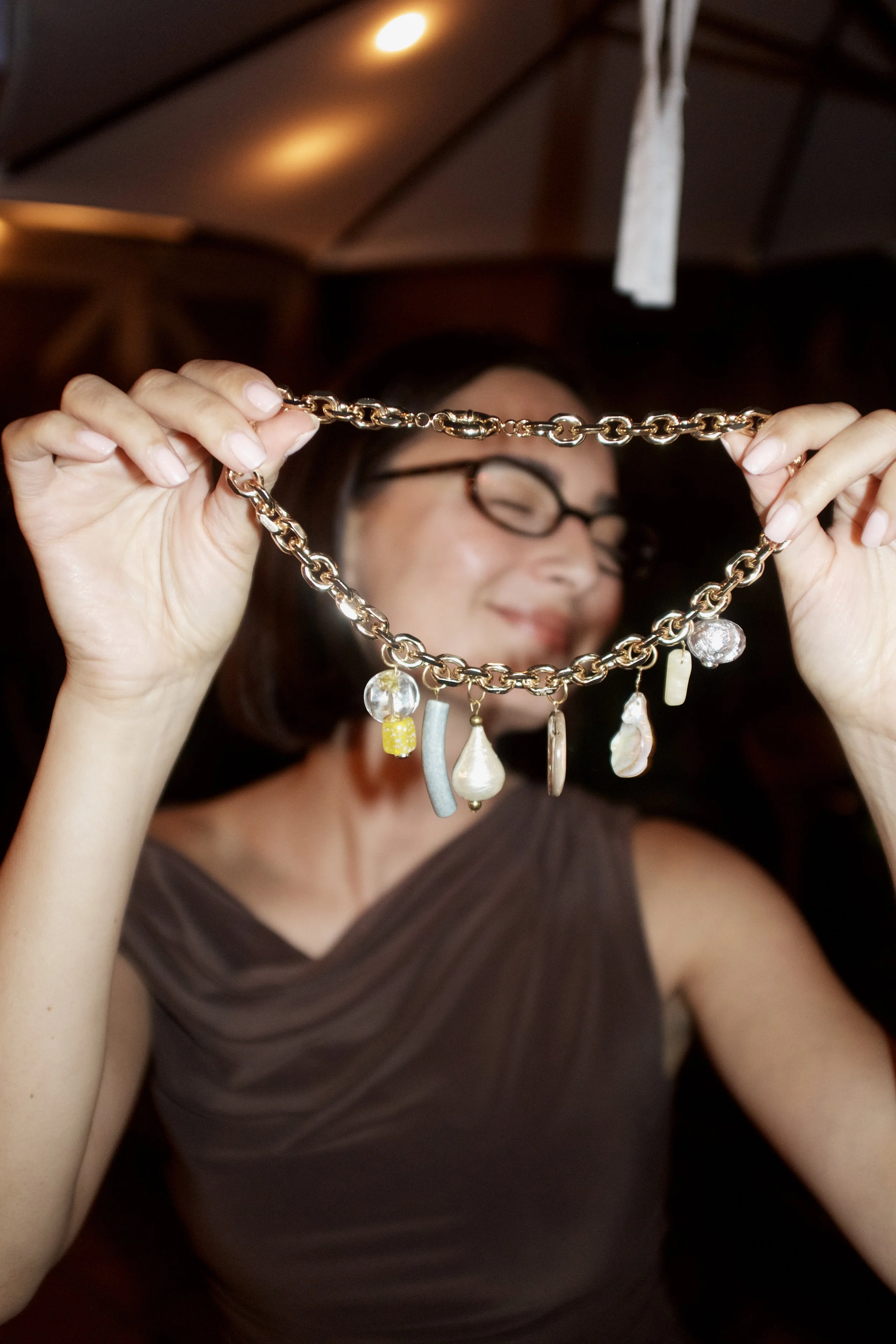 A woman wearing glasses and a brown dress is holding up a gold necklace with various charms, including pearls, shells, and beads, in a dimly lit room with a wooden ceiling.