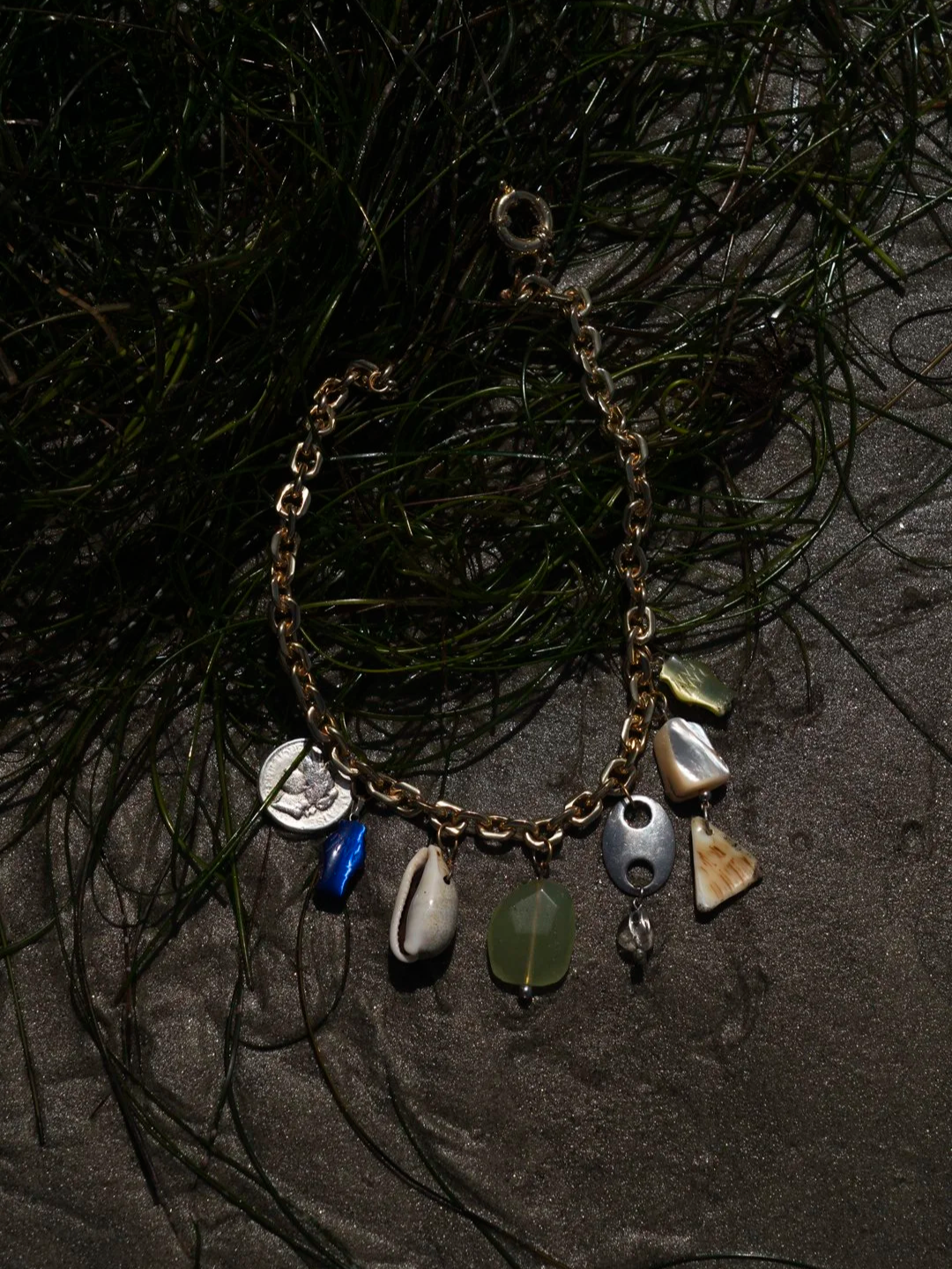 A gold chain necklace with various shell and stone charms, resting on green seaweed on a dark background.
