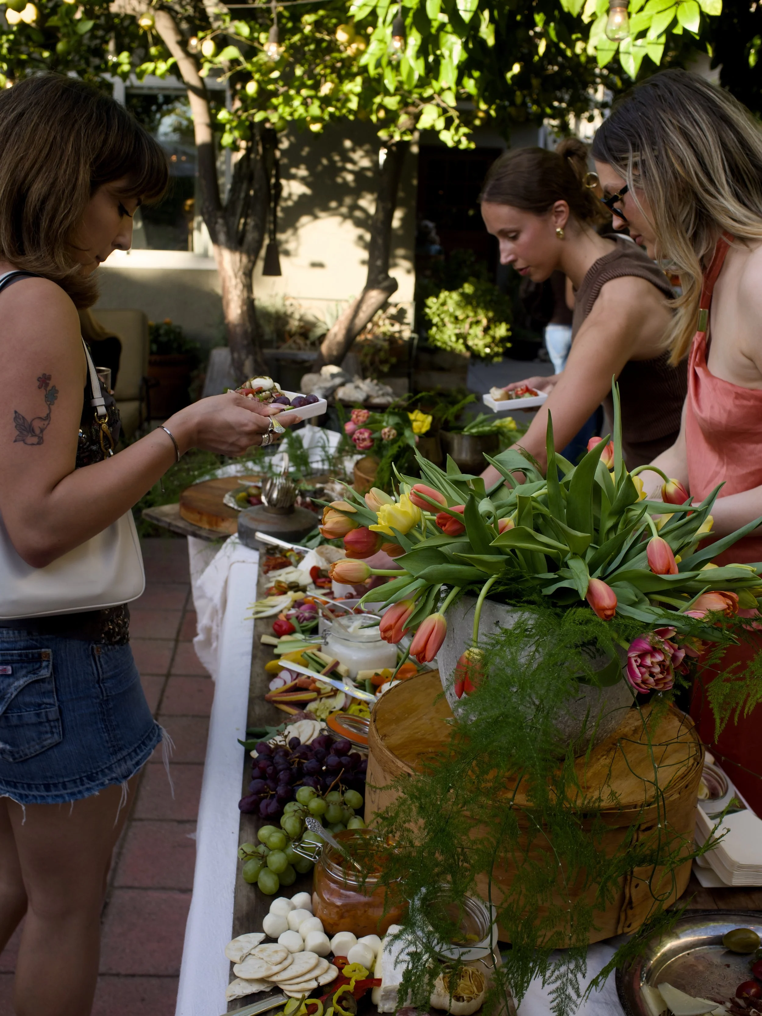 People at an outdoor picnic table with a variety of cheeses, grapes, vegetables, and flowers, under a shaded tree.