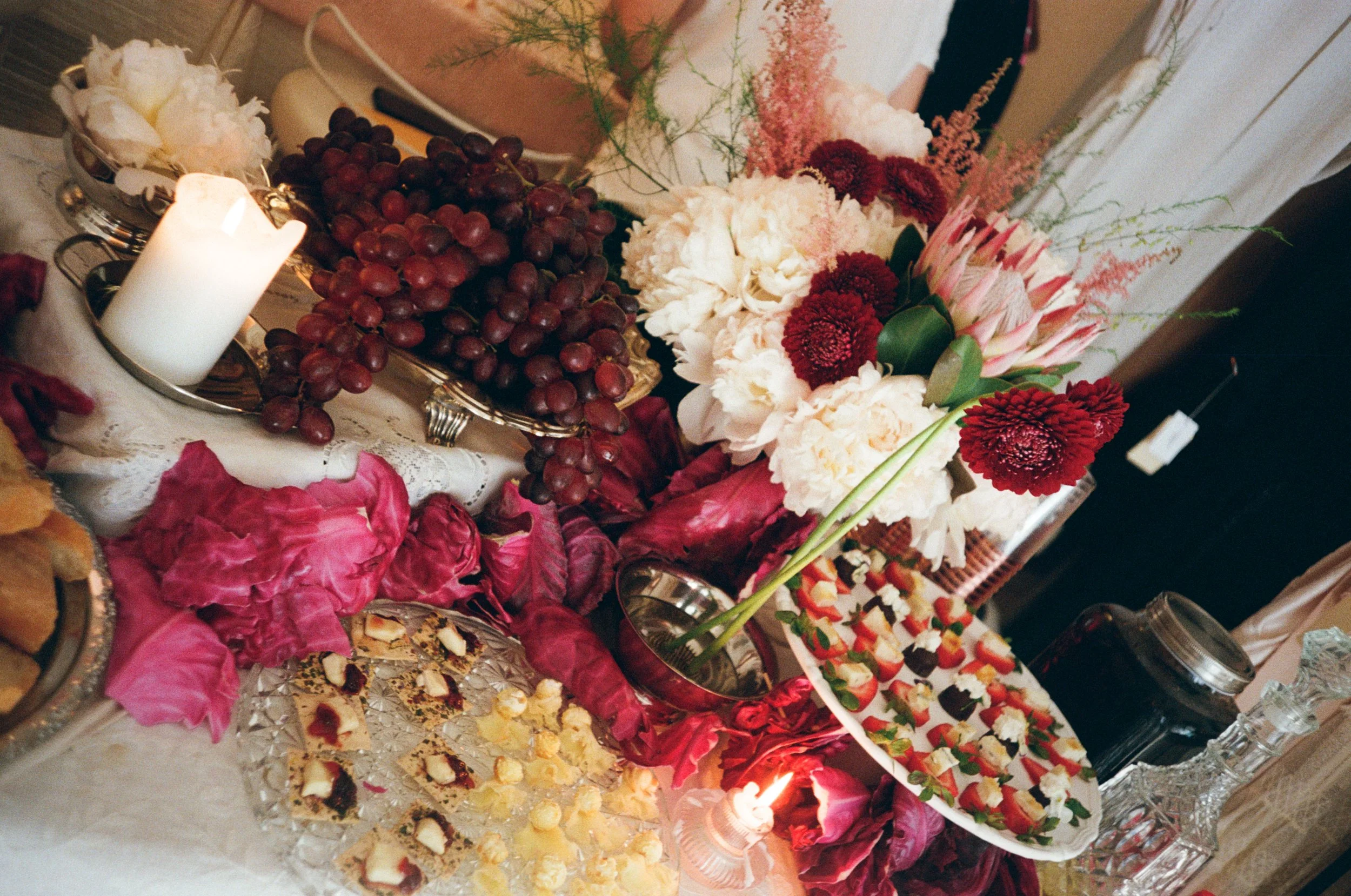 A table decorated with pink, white, and red flowers, a bunch of red grapes, a white lit candle, and various trays of desserts, including small layered treats and fruit-topped pastries.
