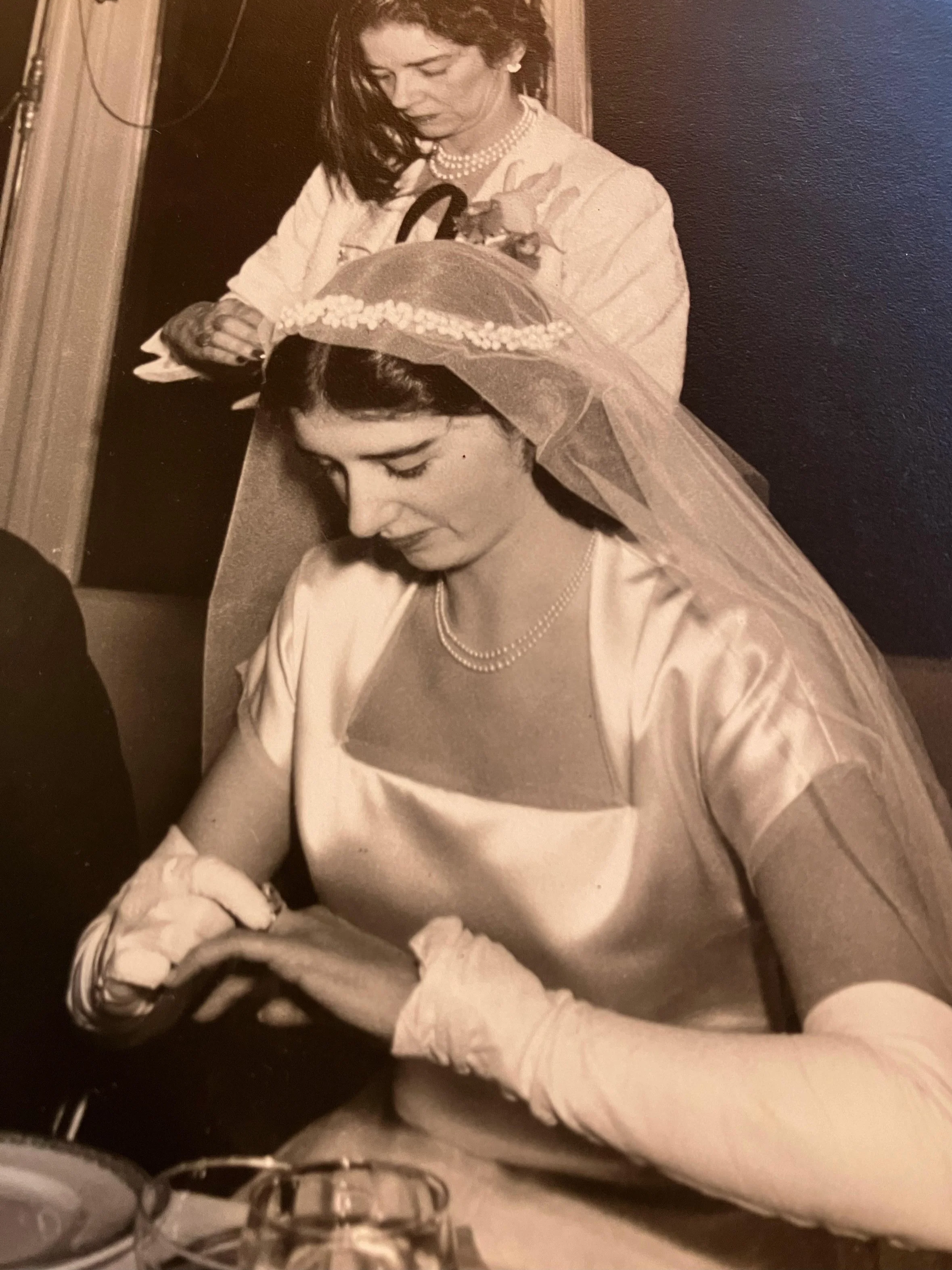 A woman wearing a wedding dress, veil, and pearl necklace is seated at a table, looking down, possibly praying or reflecting. An older woman, dressed formally, is standing behind her, adjusting something on her head or veil.