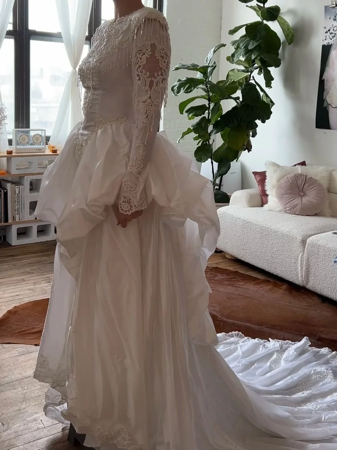 A woman in a wedding dress standing in a bright living room with a large window, green houseplant, white sofa, and decorative pillows.