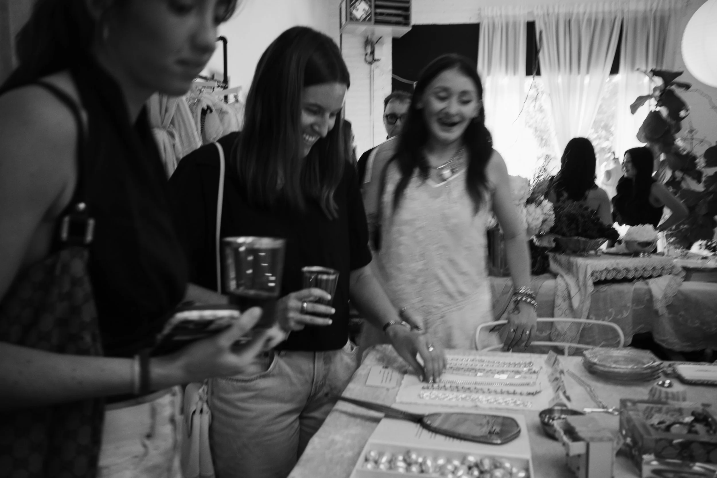 Group of women gathered around a table with jewelry, smiling and looking at items, in a cheerful indoor setting.