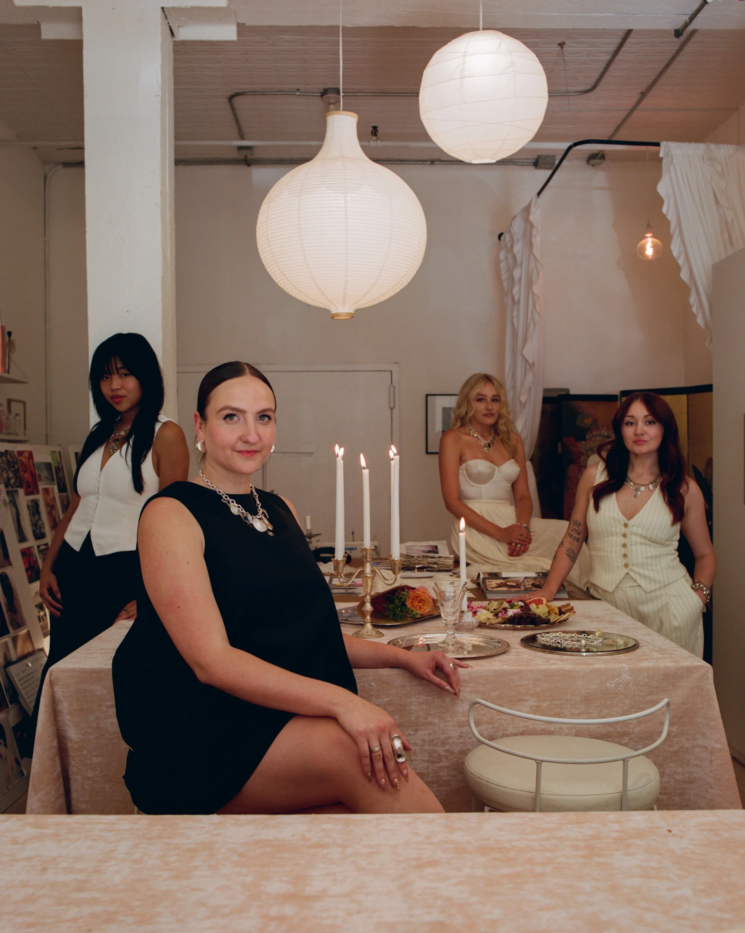 Four women in elegant dresses gathered around a dining table with candles and floral arrangements, in a warmly lit room with paper lantern ceiling lights.
