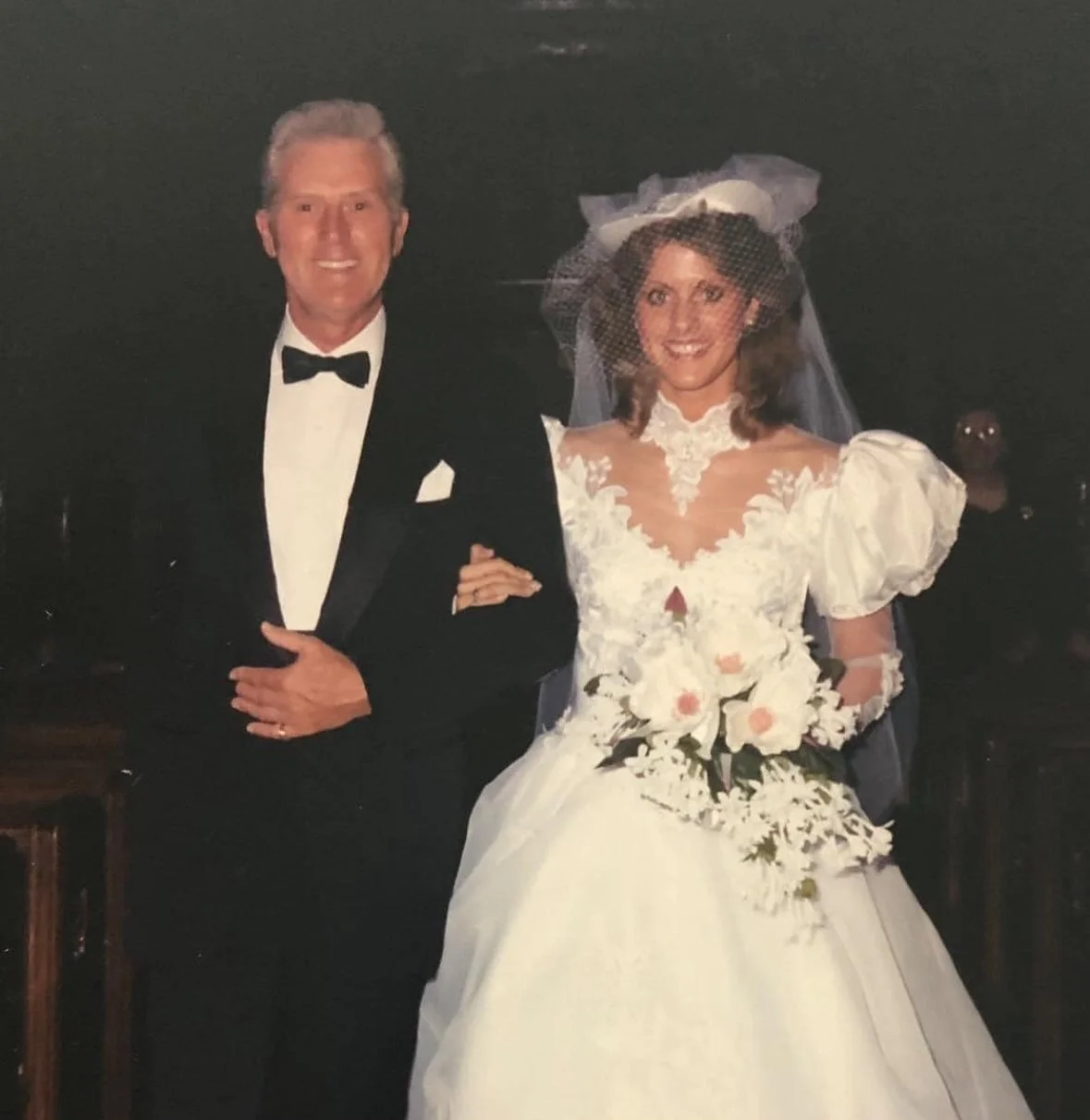 A man in a tuxedo and a woman in a wedding dress standing together, smiling at a wedding or formal event.