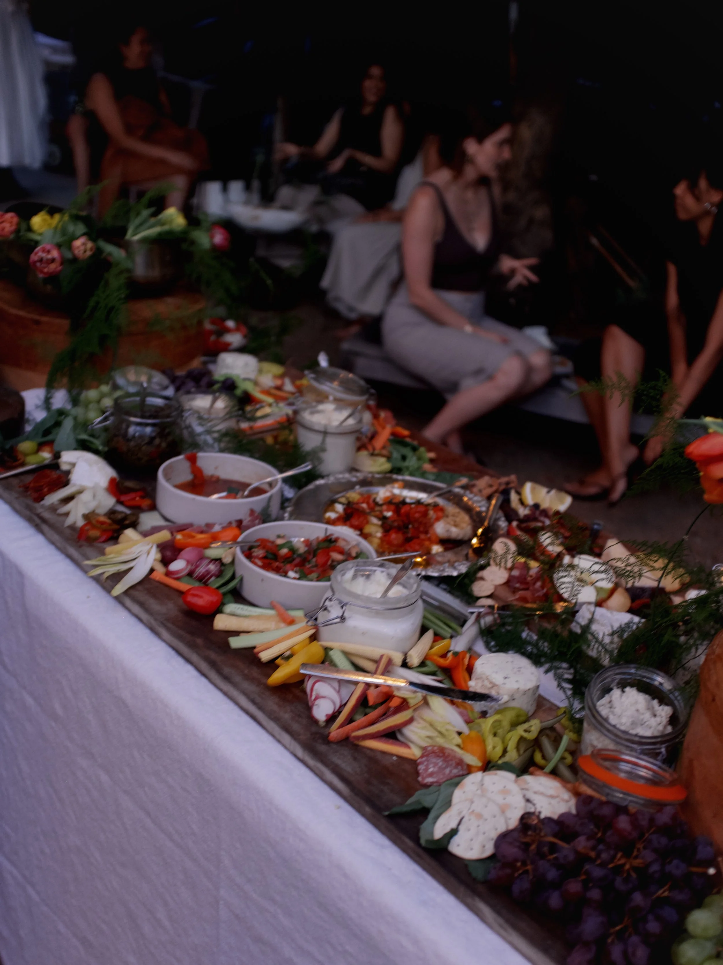 A buffet table with various vegetables, cheeses, and appetizers, decorated with greenery; people seated in the background at a social gathering.