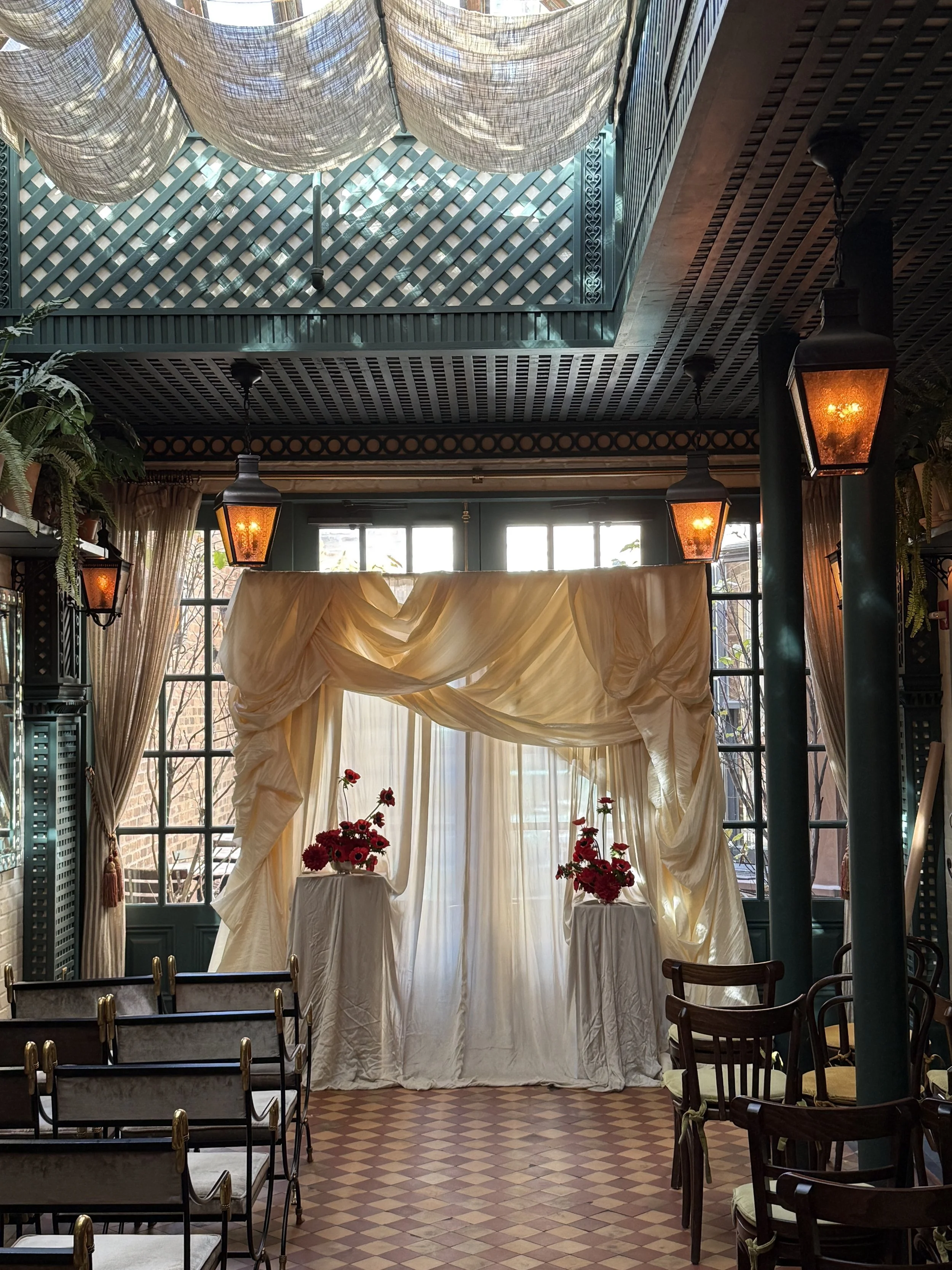 Intimate indoor wedding altar with cream drapery and red flowers, set in a decorated room with large windows, wooden chairs, and hanging lanterns.
