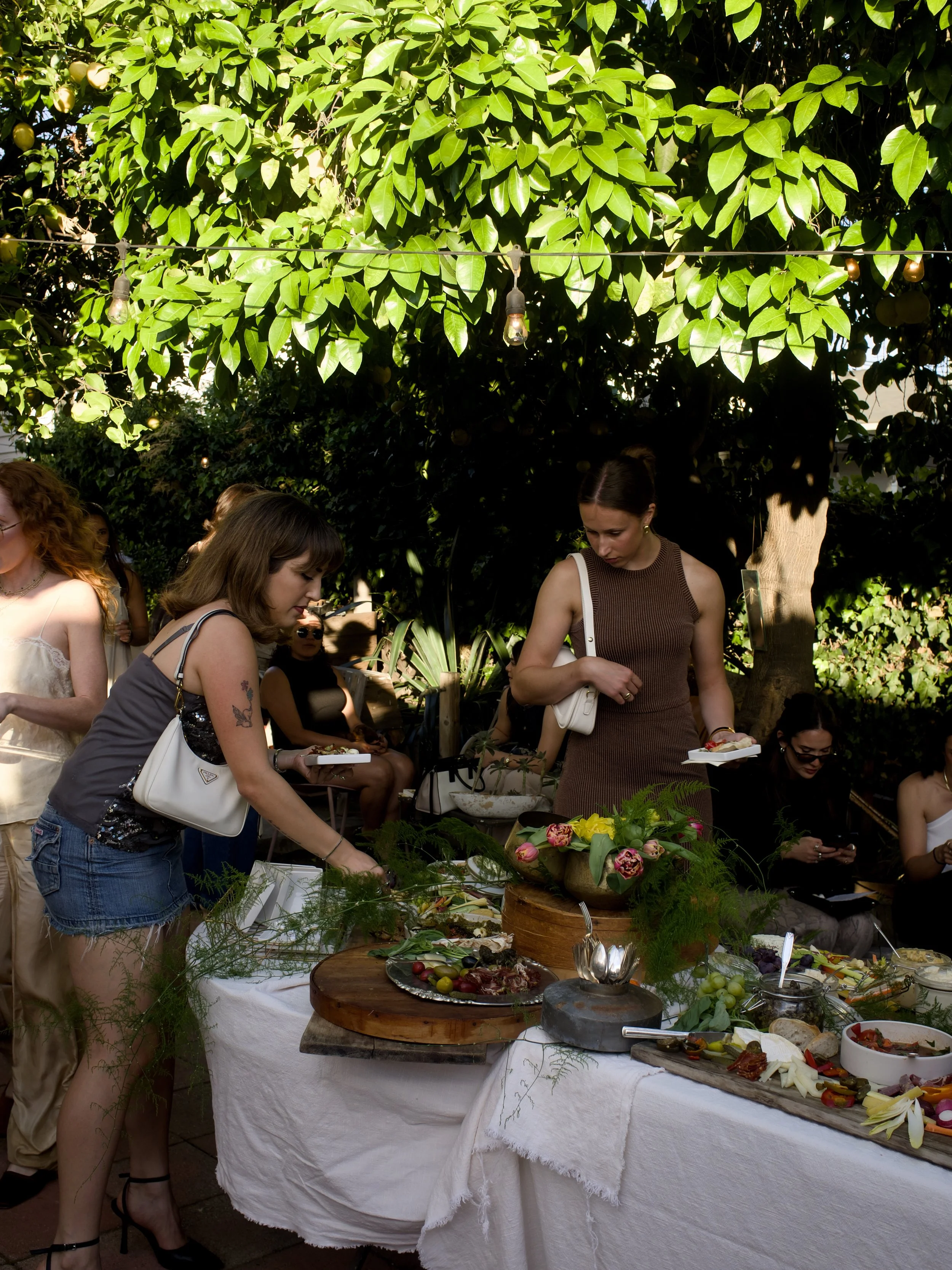 People gathered around a table outdoors with various dishes, greenery, and trees overhead.