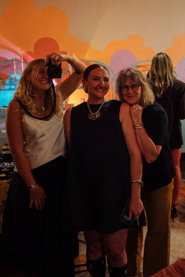 Three women standing together in a colorful, decorated room, smiling at the camera, taking a selfie.