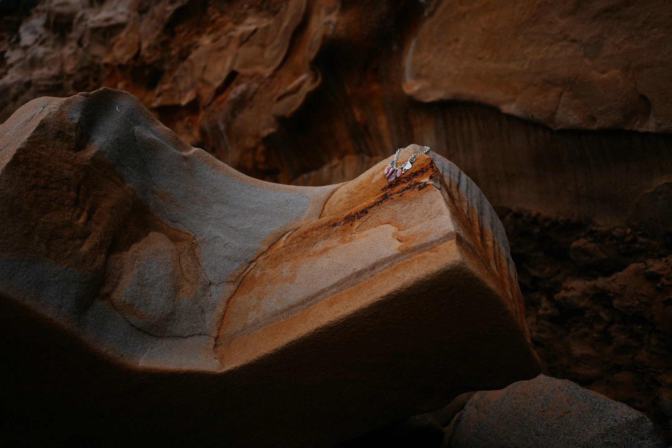 A rocky cave interior with shadows cast on the uneven surface and a silver necklace with pink beads resting on a large rock.