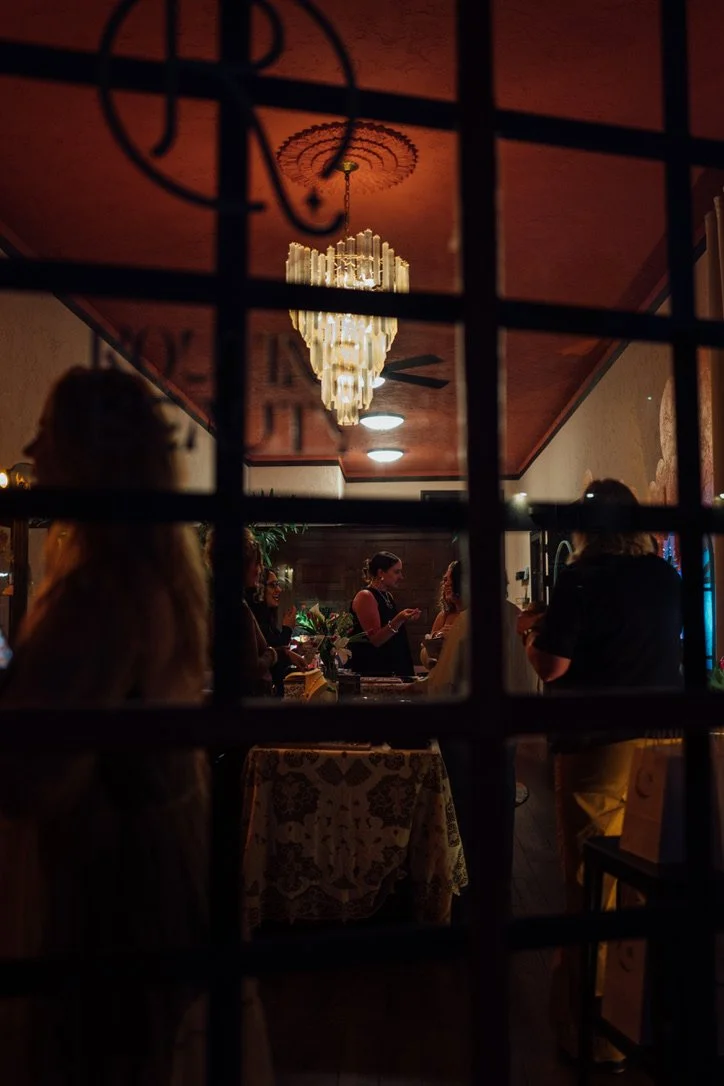 A cozy indoor restaurant scene seen through a window, illuminated by a grand chandelier, with several people gathered around a table, some standing and some looking at their phones.