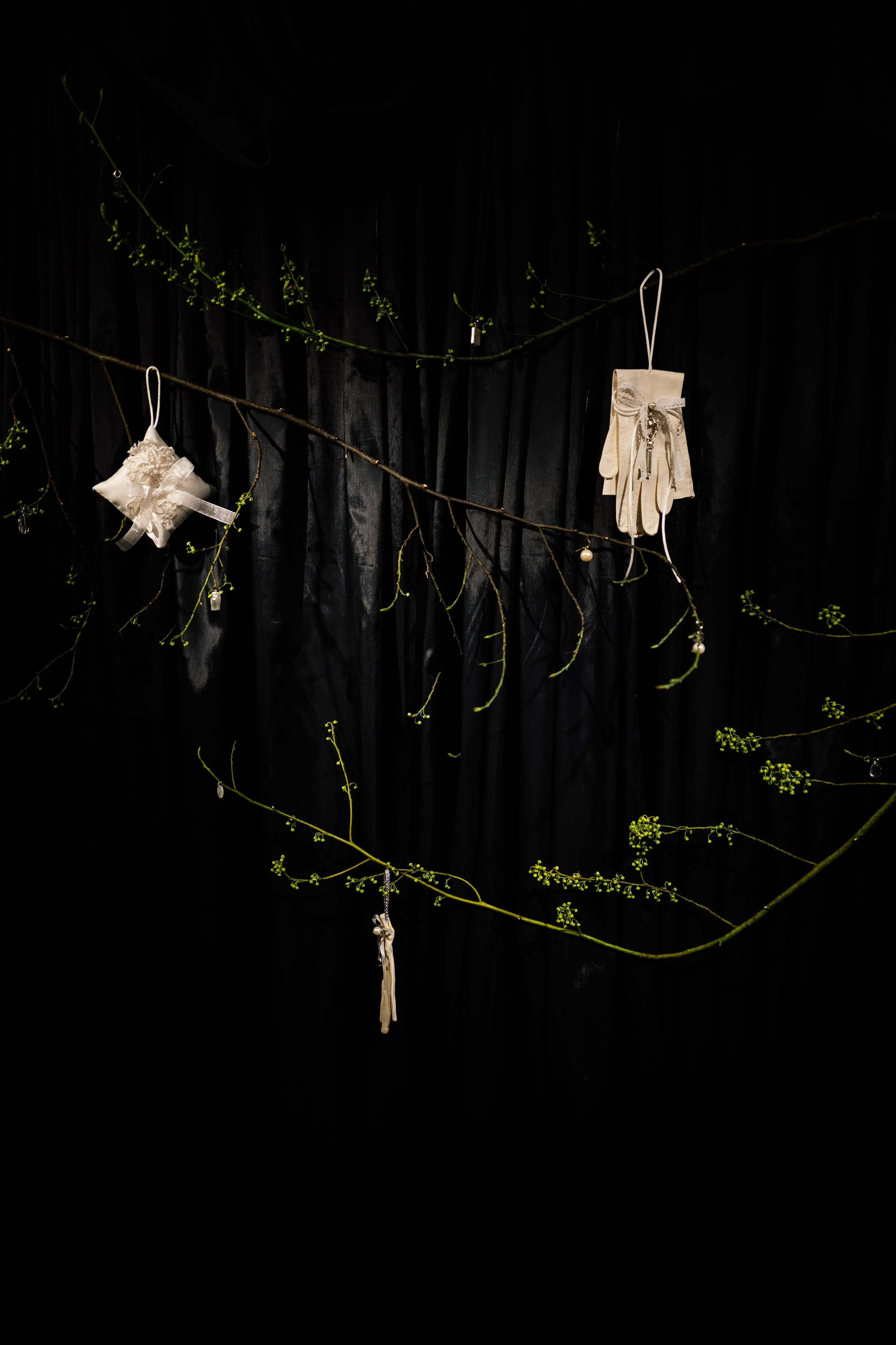 Decorative display featuring hanging cream-colored gloves, a small pillow, and a ribbon on thin branches with small green leaves, against a black background.