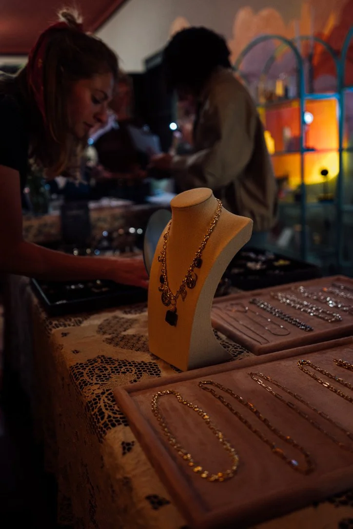 Jewelry display table with gold necklaces and bracelets, with two women browsing jewelry in a dimly lit, colorful indoor setting