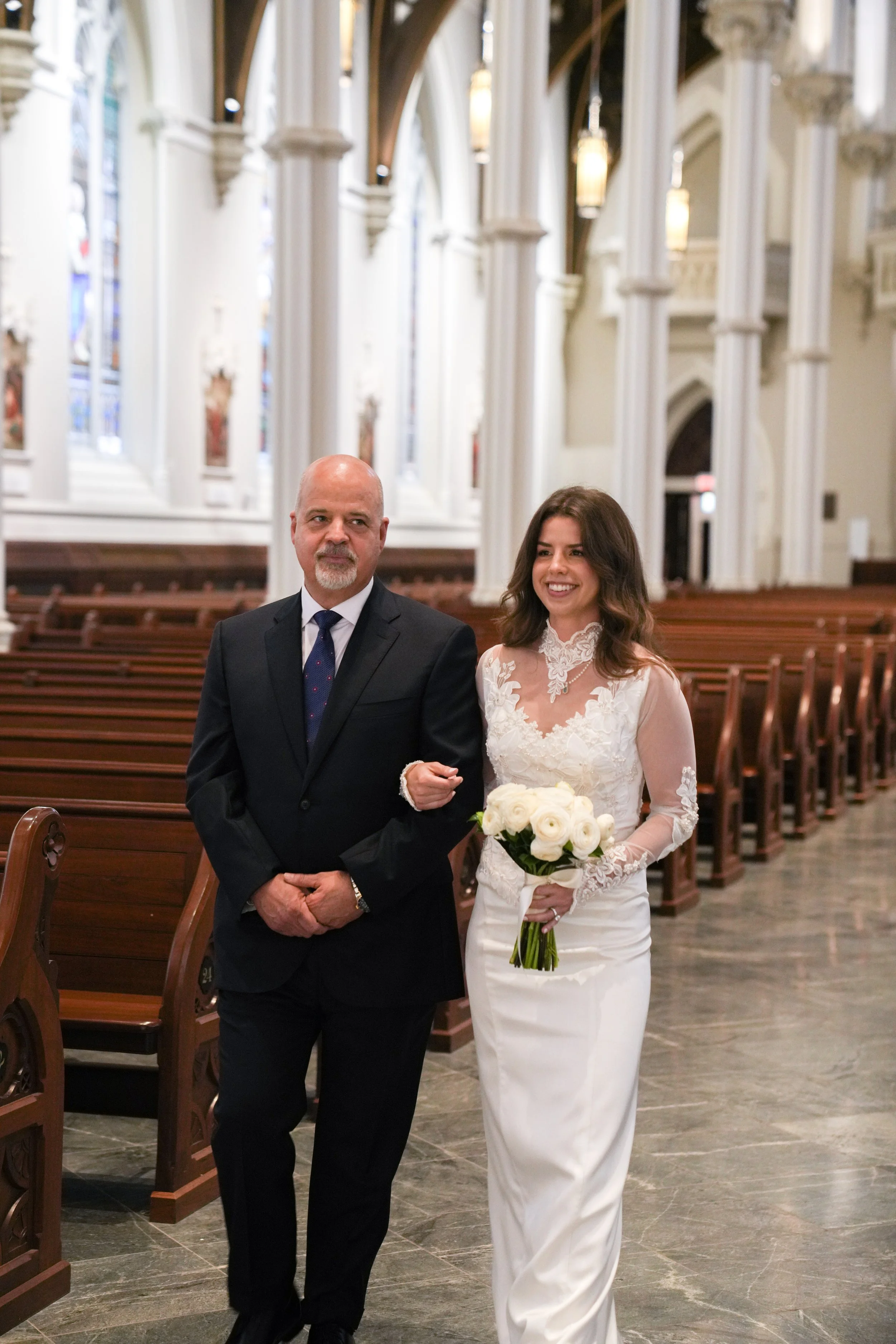 A bride in a white wedding dress holding a bouquet of white roses stands next to an older man in a black suit inside a church with tall white columns and stained glass windows.