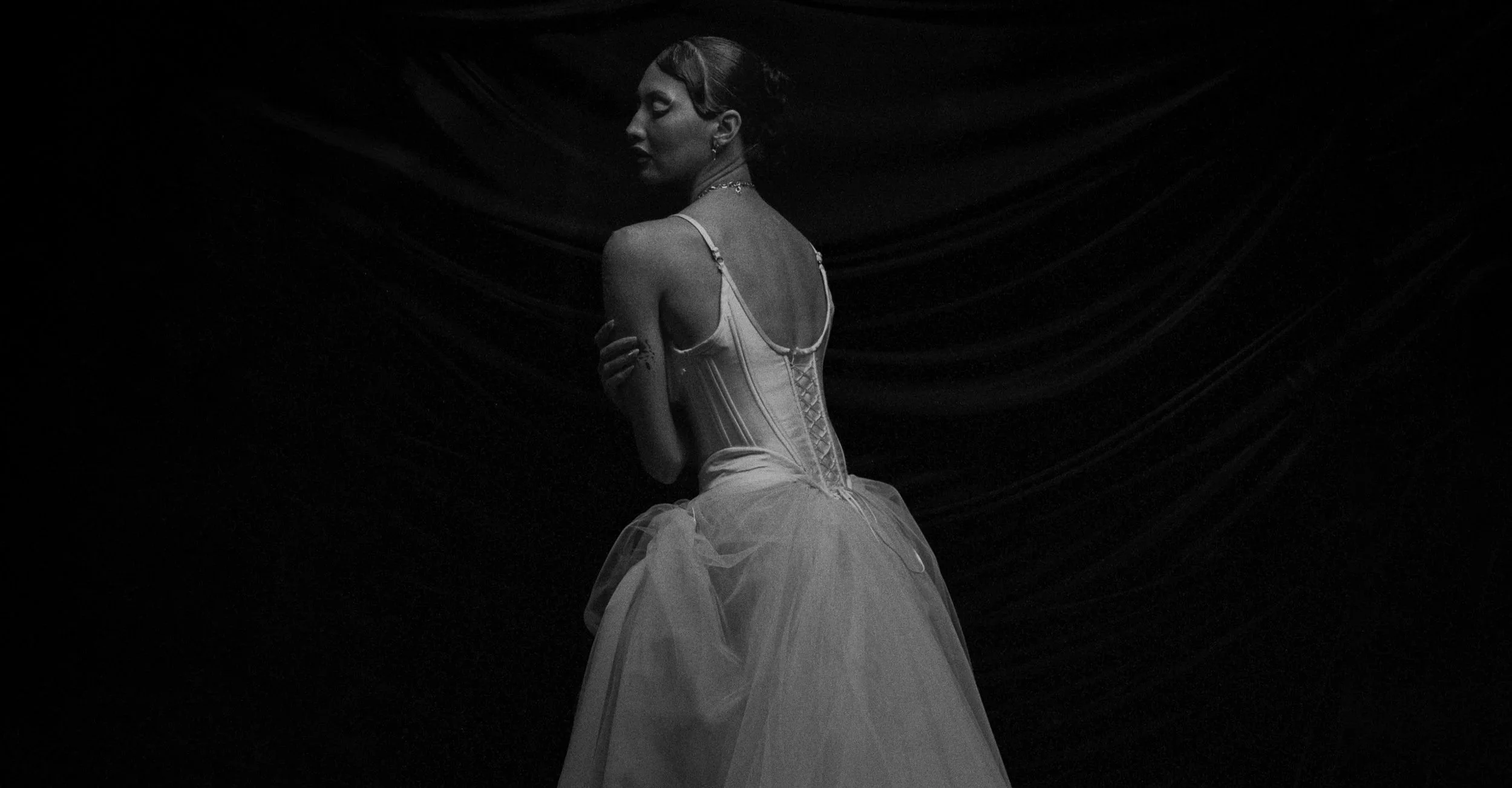 A black and white photograph of a woman in a vintage wedding dress, sitting with her back turned, arms crossed, on a dark background with draped fabric.
