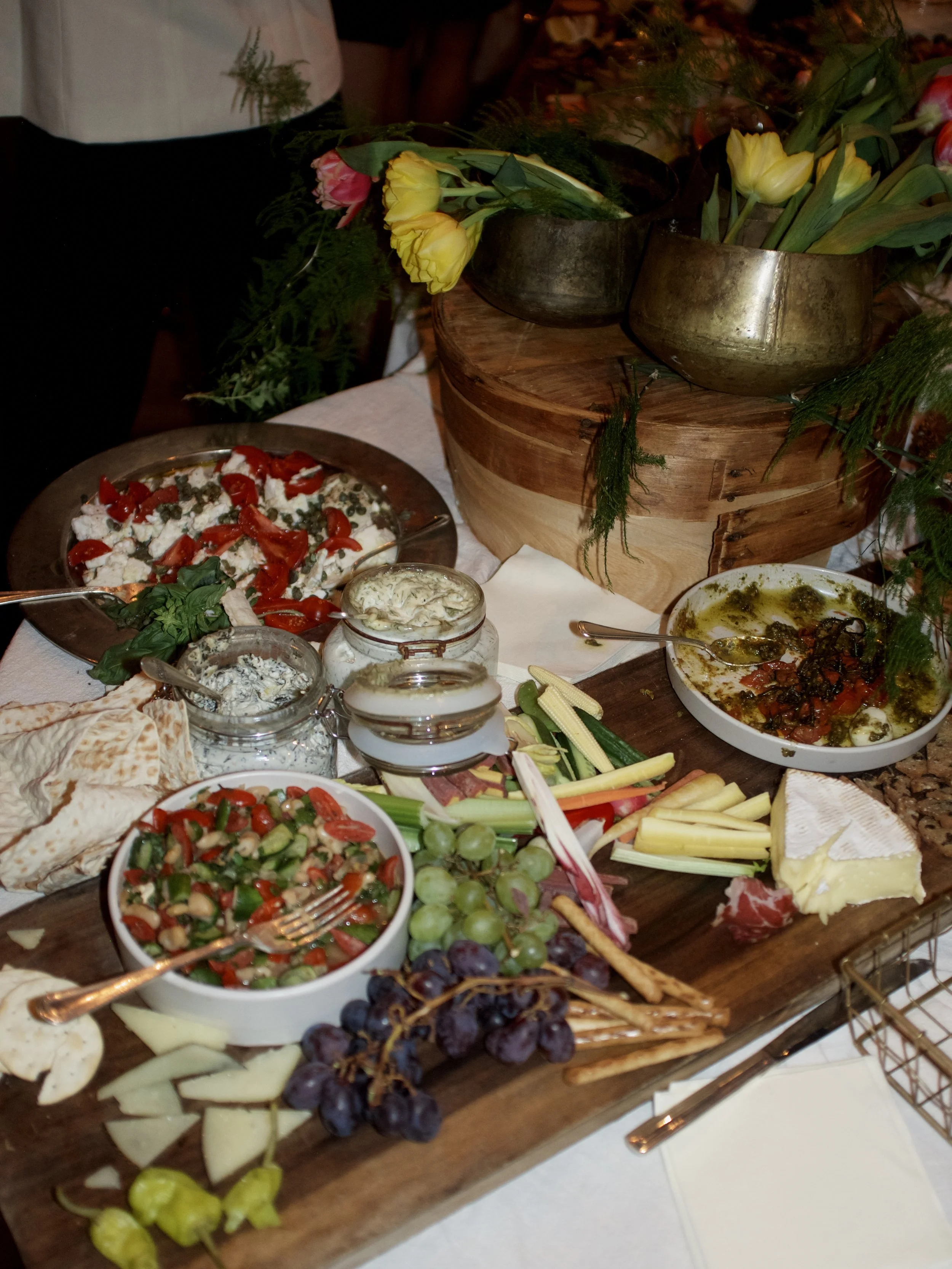 Arrangement of assorted foods, cheeses, grapes, vegetables, and salads on a wooden table, with a centerpiece of flower vases and greenery.