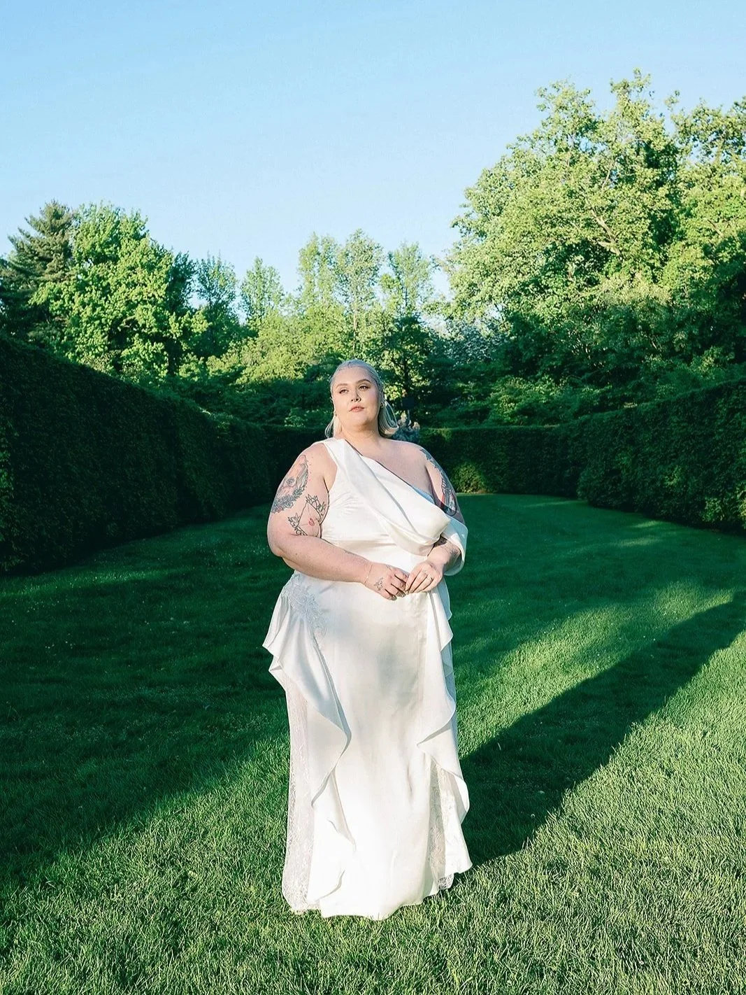 A woman in a white dress standing on a well-manicured lawn with trees and bushy greenery in the background during daytime.