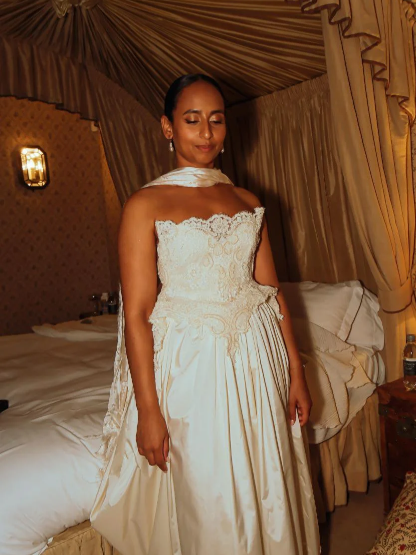 A woman in an elegant white lace strapless gown with intricate embroidery, standing in a warmly lit, ornate bedroom with draped curtains and a bed with white bedding.