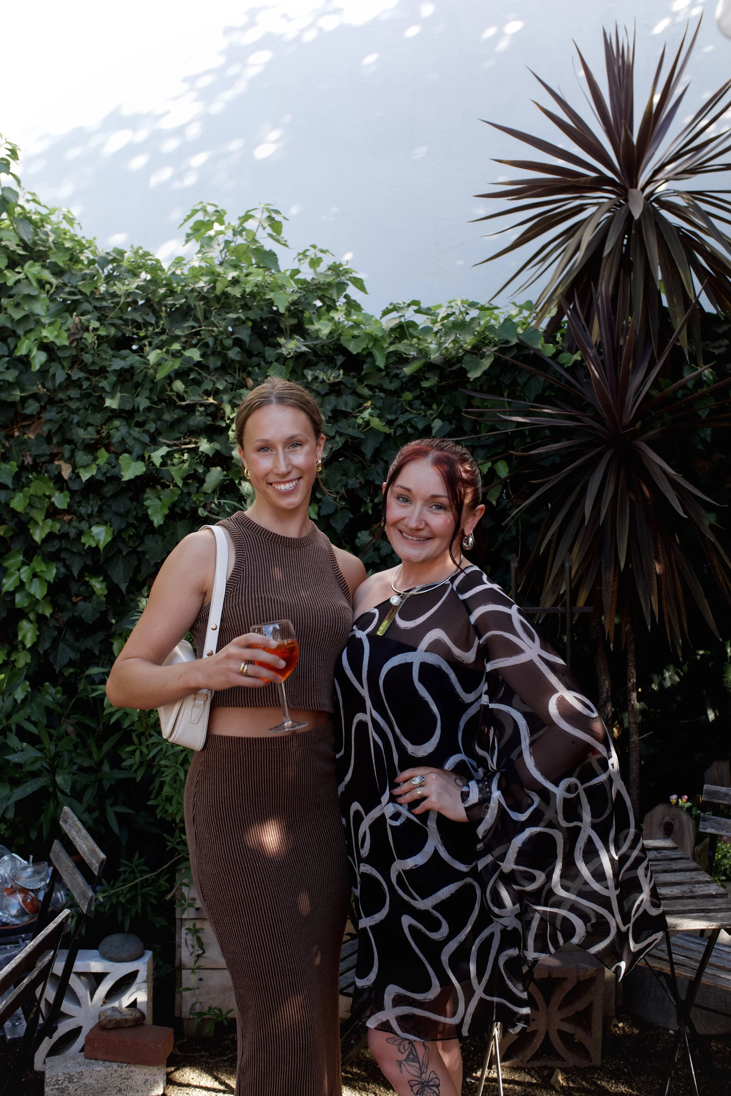 Two women standing outdoors in a garden, smiling at the camera. One holds a glass of red drink, and they are surrounded by green foliage and large potted plants.
