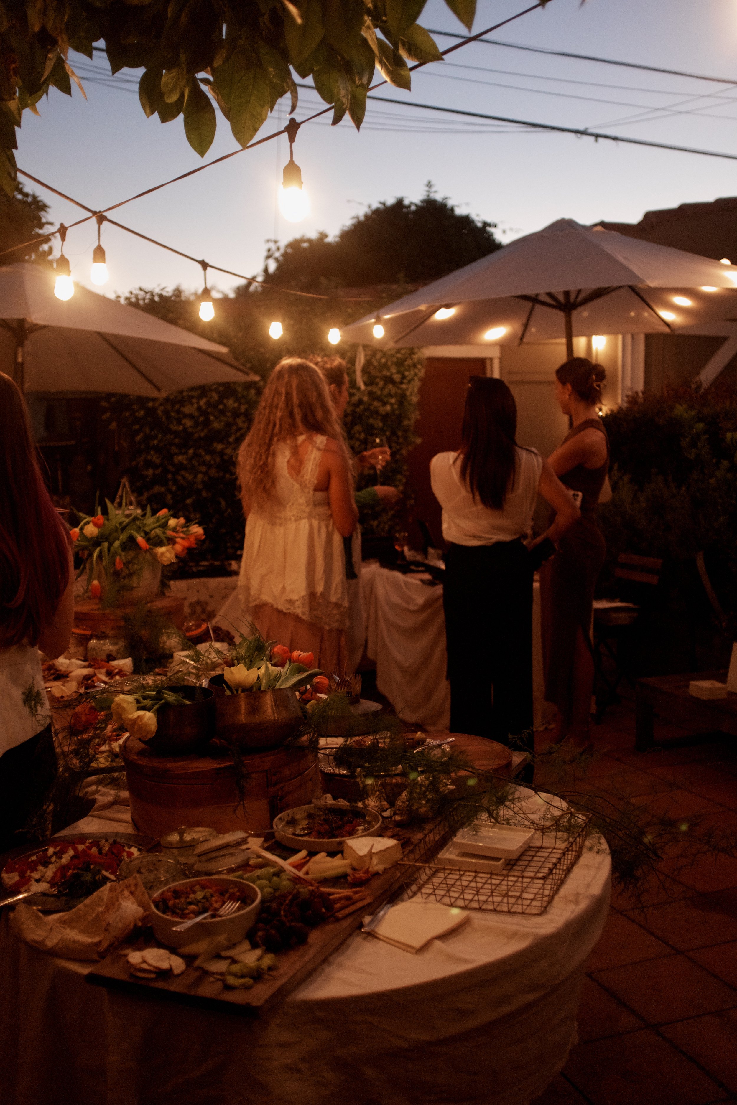 People socializing at an outdoor evening gathering with string lights, umbrellas, and a table filled with various foods and flowers.