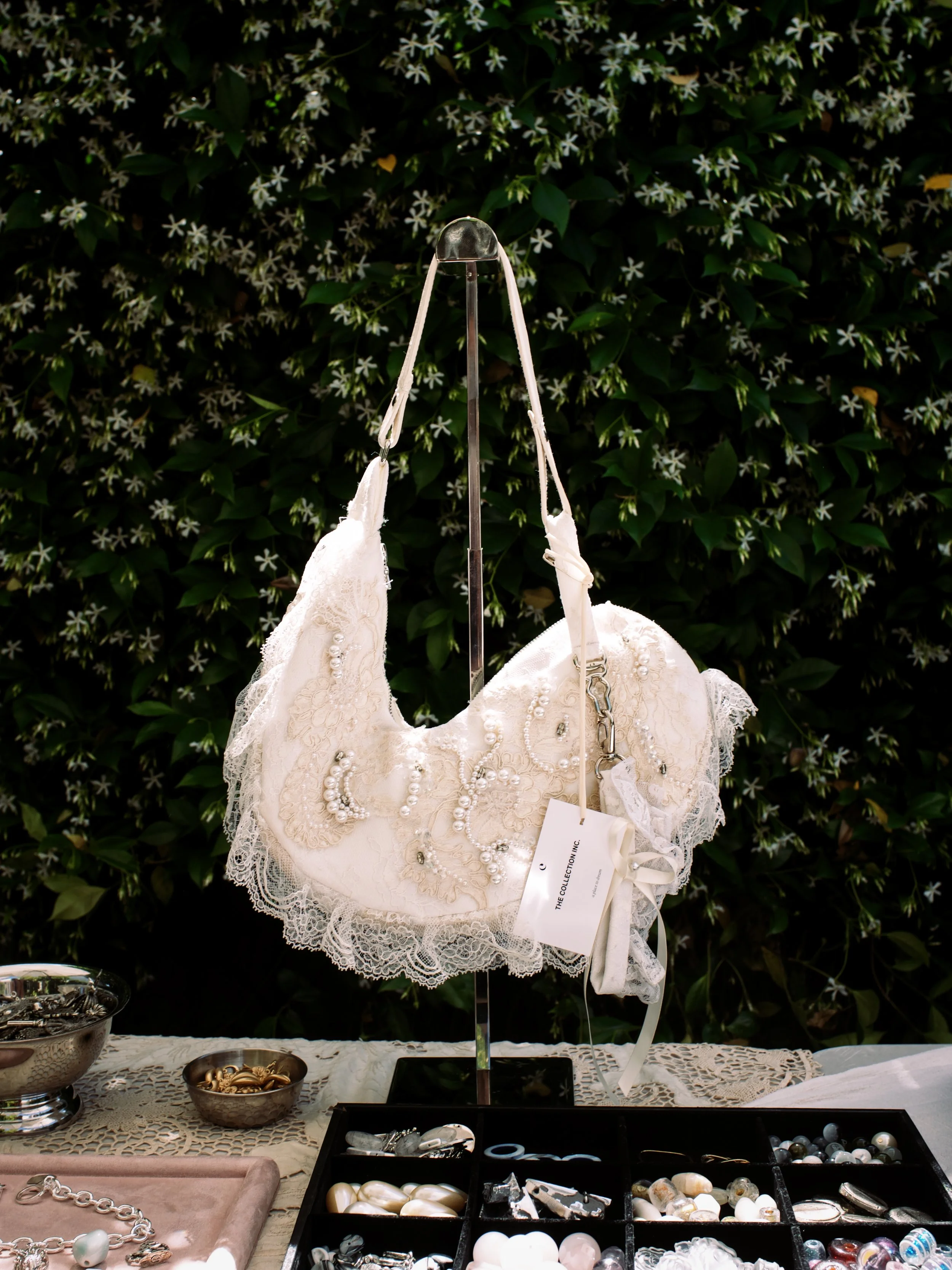Table display of jewelry including a dress form with pearl and lace clothing, bowls filled with jewelry pieces, and various jewelry accessories on a table with a lace tablecloth in front of a green leafy background.