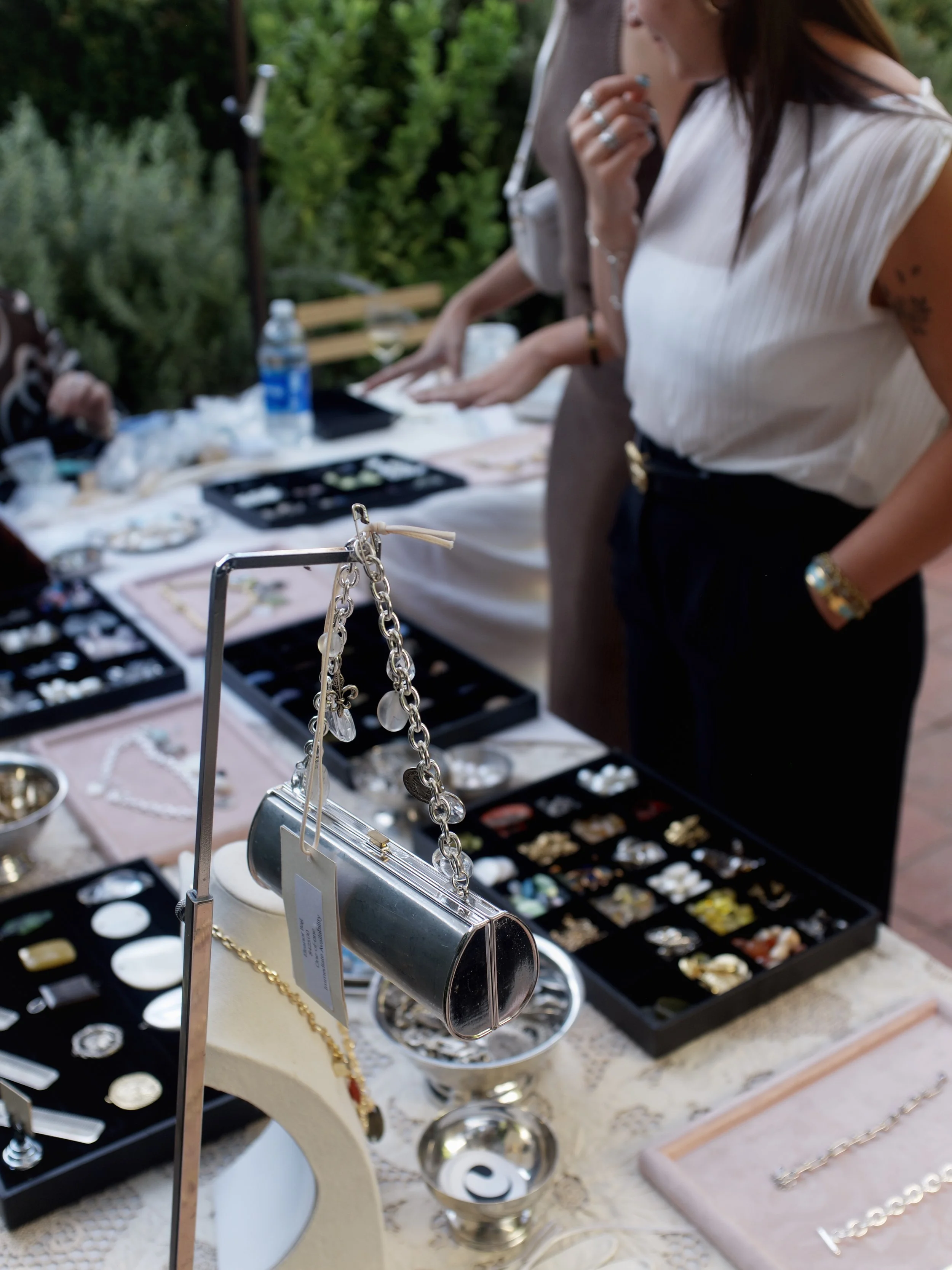 Jewelry display table with necklaces, earrings, and bracelets at an outdoor market, with blurred greenery in the background.