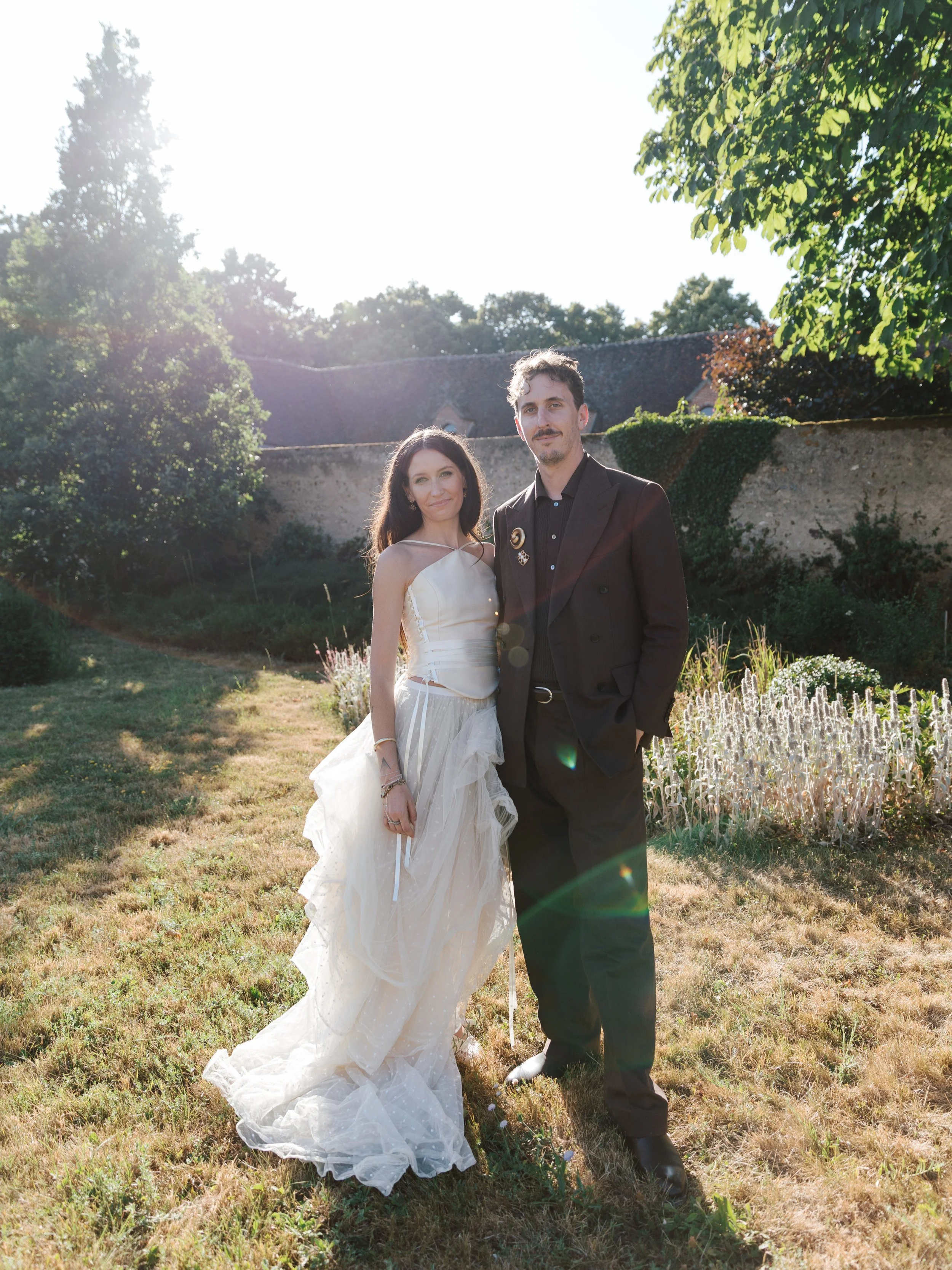 A couple standing outdoors in a garden during late afternoon sunlight, with a stone wall and trees in the background. The woman is wearing a white dress and the man a dark suit.