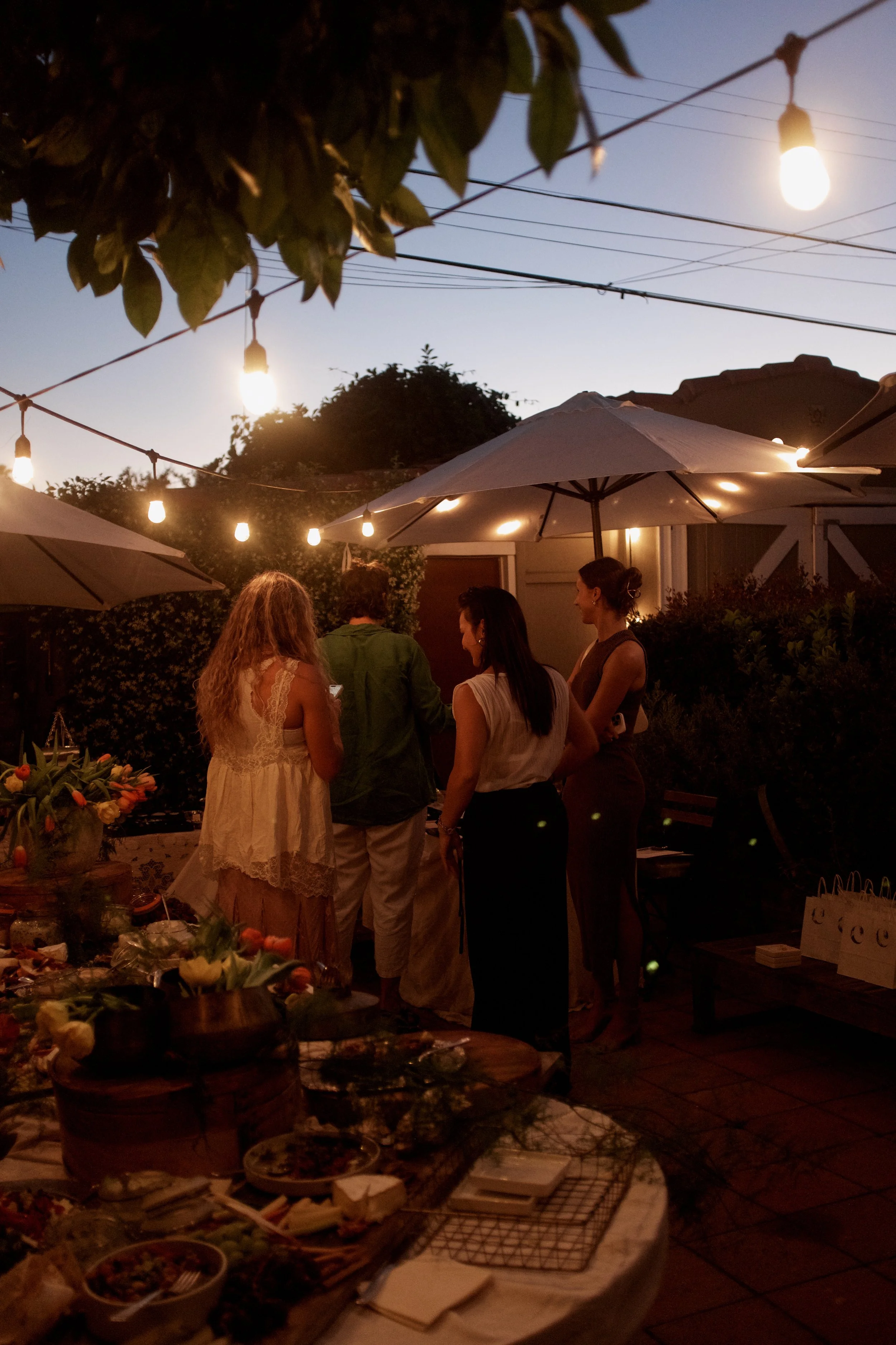 People socializing at a nighttime outdoor gathering with string lights and umbrellas, surrounded by tables with food and drinks.
