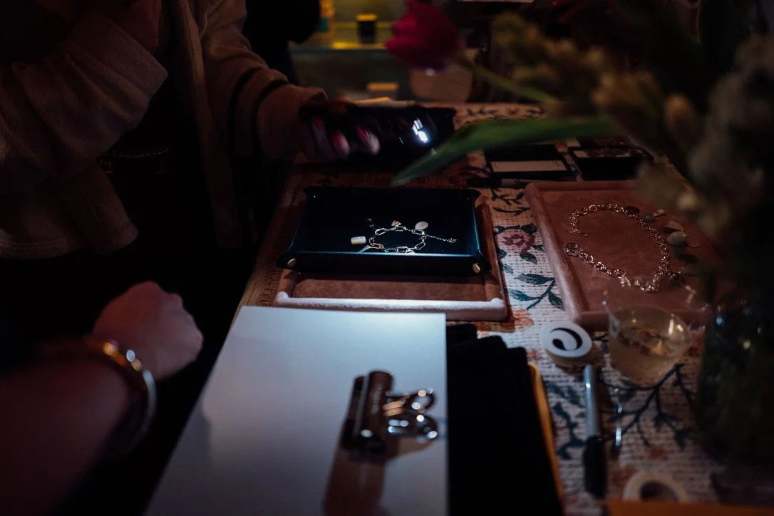Jewelry display on a floral tablecloth with some people in background and a drink on the side.