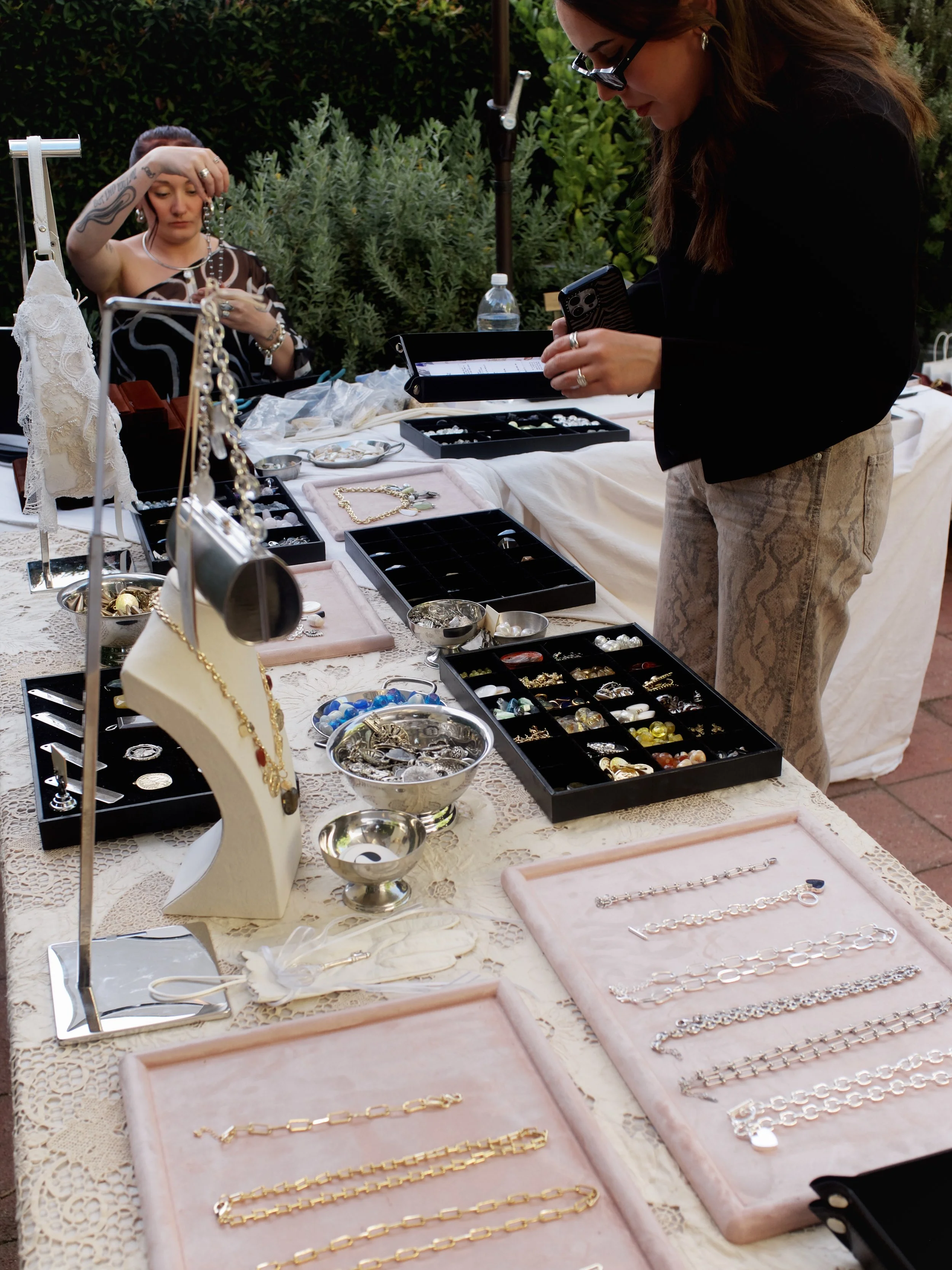 Jewelry displayed on a table at an outdoor market, with two women browsing and examining necklaces, bracelets, and earrings surrounded by green foliage.