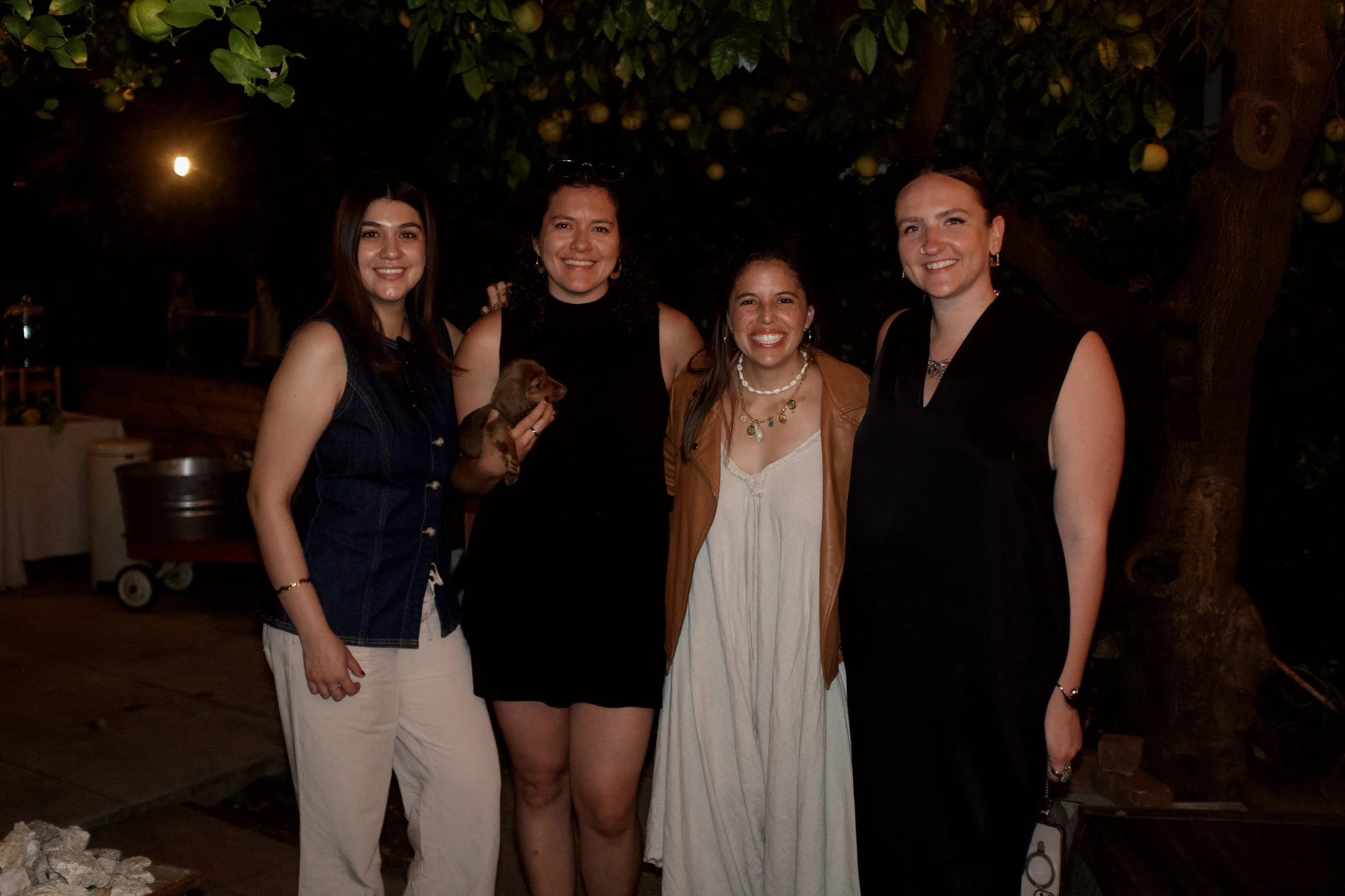 Four women smiling and standing close together outdoors at night under a tree with yellow fruit. One woman is holding a small puppy. They appear to be at a casual gathering or party.