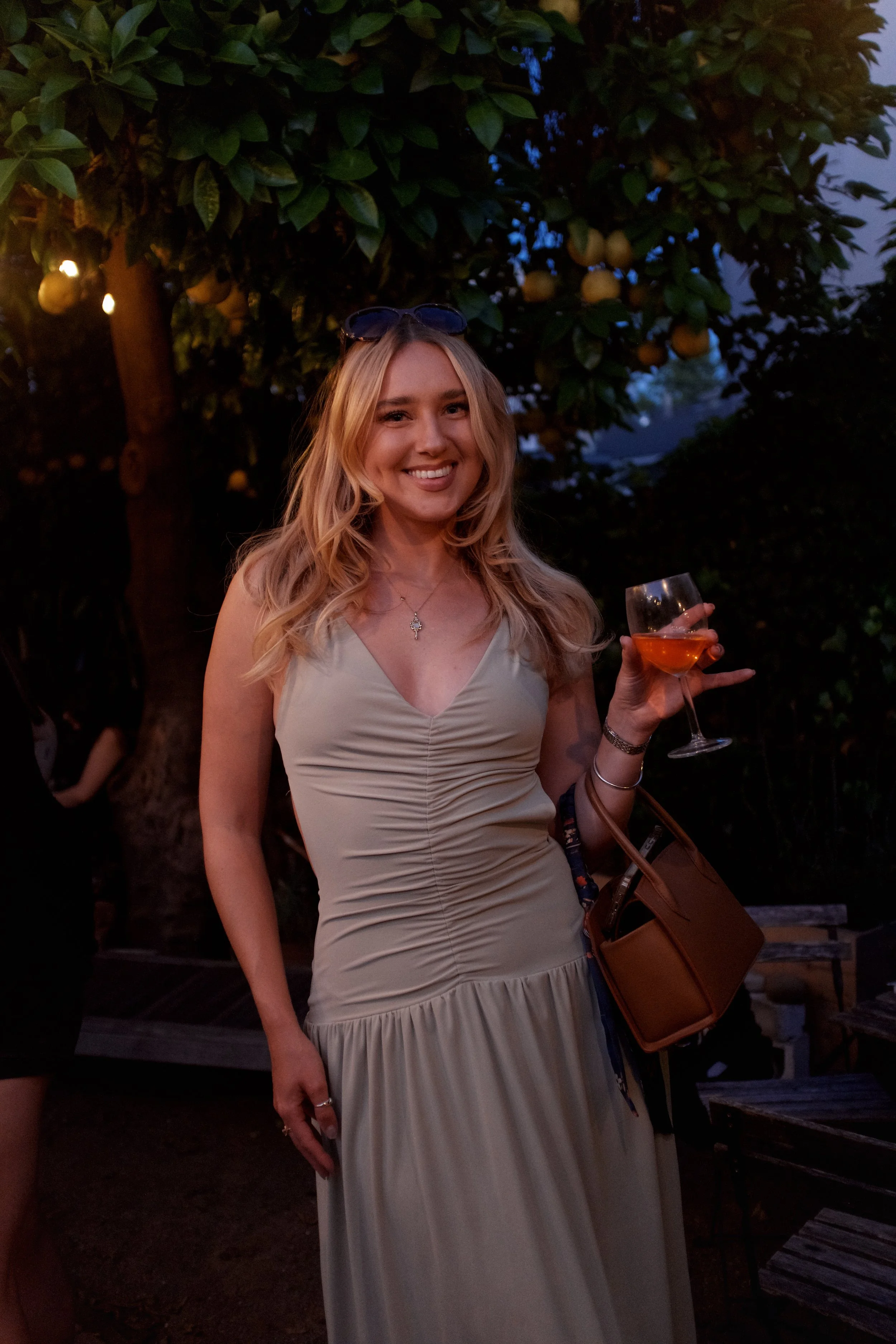 A woman with blonde hair in a light beige dress, holding a glass of rosé wine, standing outdoors near a tree with yellow fruit, during dusk or early evening.