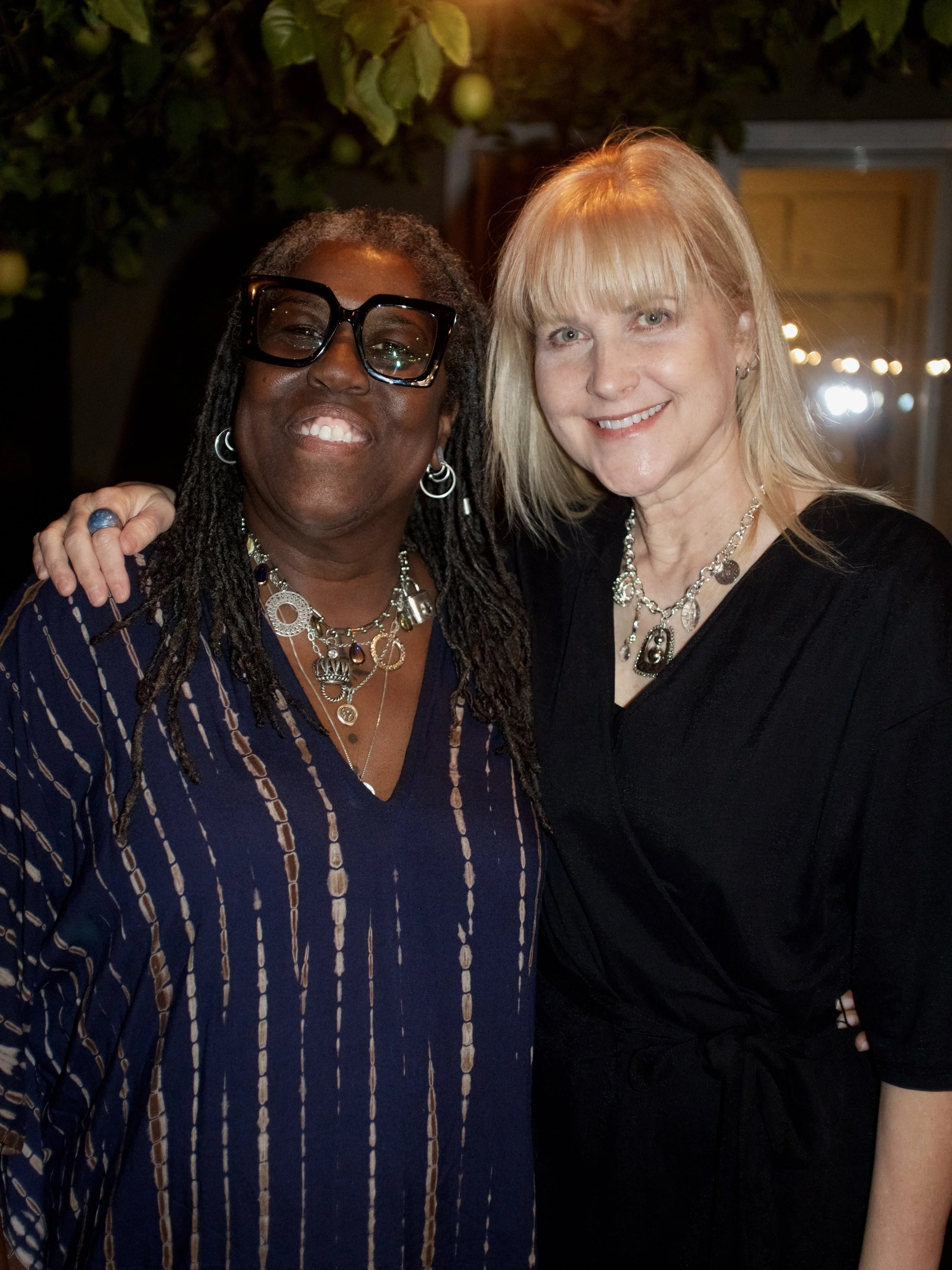 Two women with happy expressions, one with dark skin and dreadlocks wearing glasses and layered jewelry, the other with fair skin and blonde hair, standing close together, outdoors at night with string lights and greenery in the background.