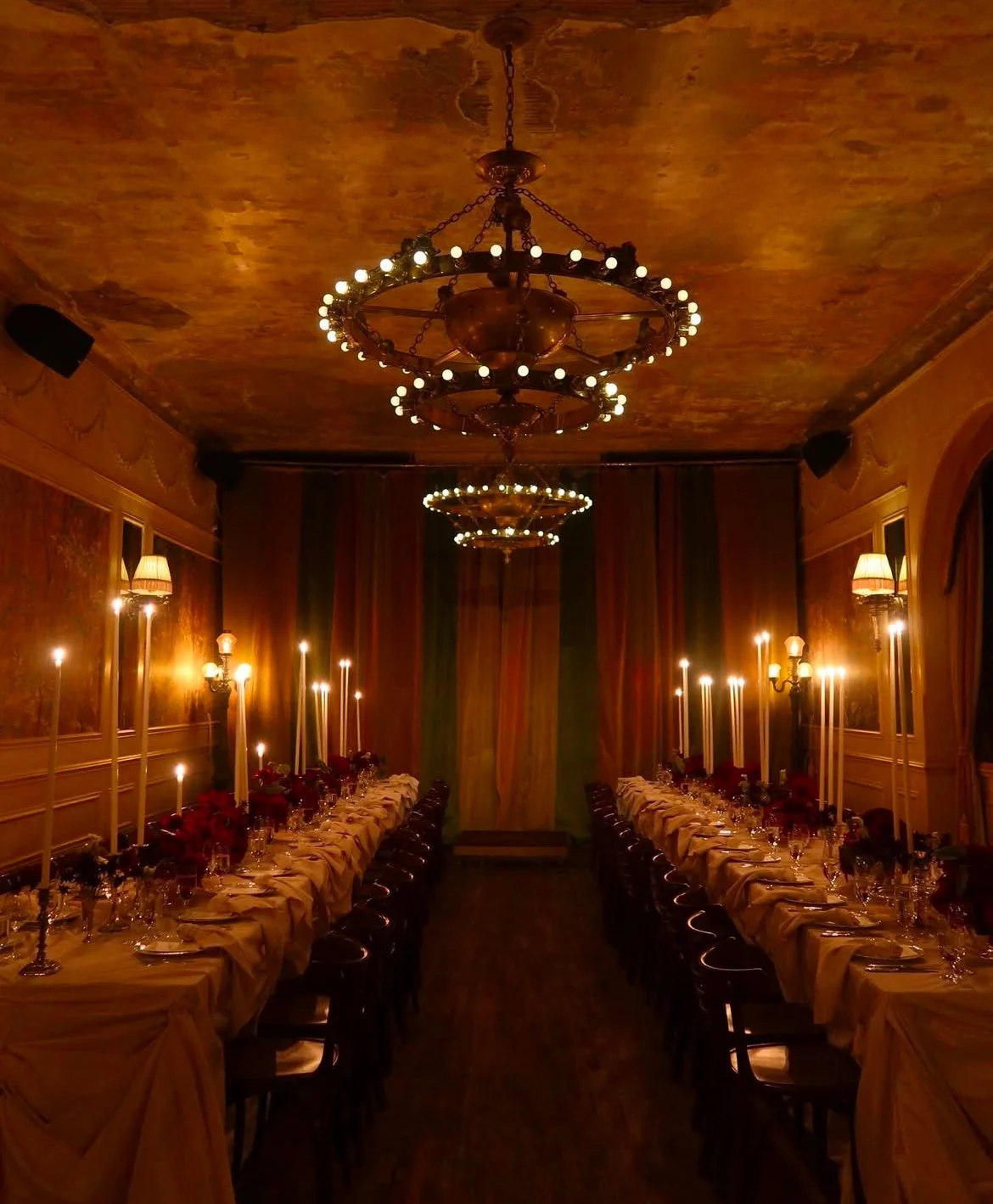 An elegantly decorated dining room with long tables set for a formal dinner, featuring candlelit centerpieces, draped tablecloths, and ornate chandeliers hanging from a richly textured ceiling.