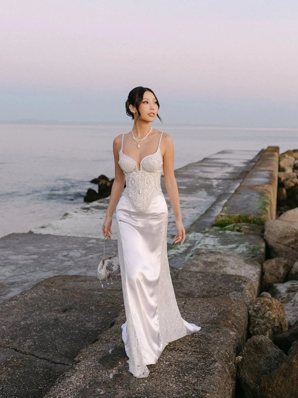 A woman in a white wedding dress walks on a rock jetty by the ocean at sunset, holding a small purse and wearing jewelry.