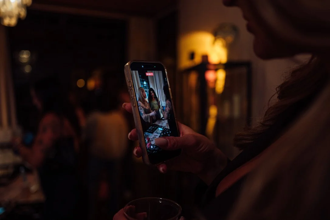 A woman is taking a photo or recording a video with her smartphone in a dimly lit social gathering or bar. The screen of the phone shows a group of people sitting at a table.
