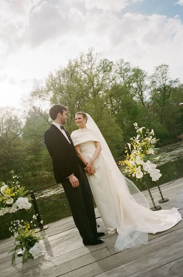 Bride and groom holding hands and smiling on outdoor wedding altar near river, with flower arrangements and trees in the background.
