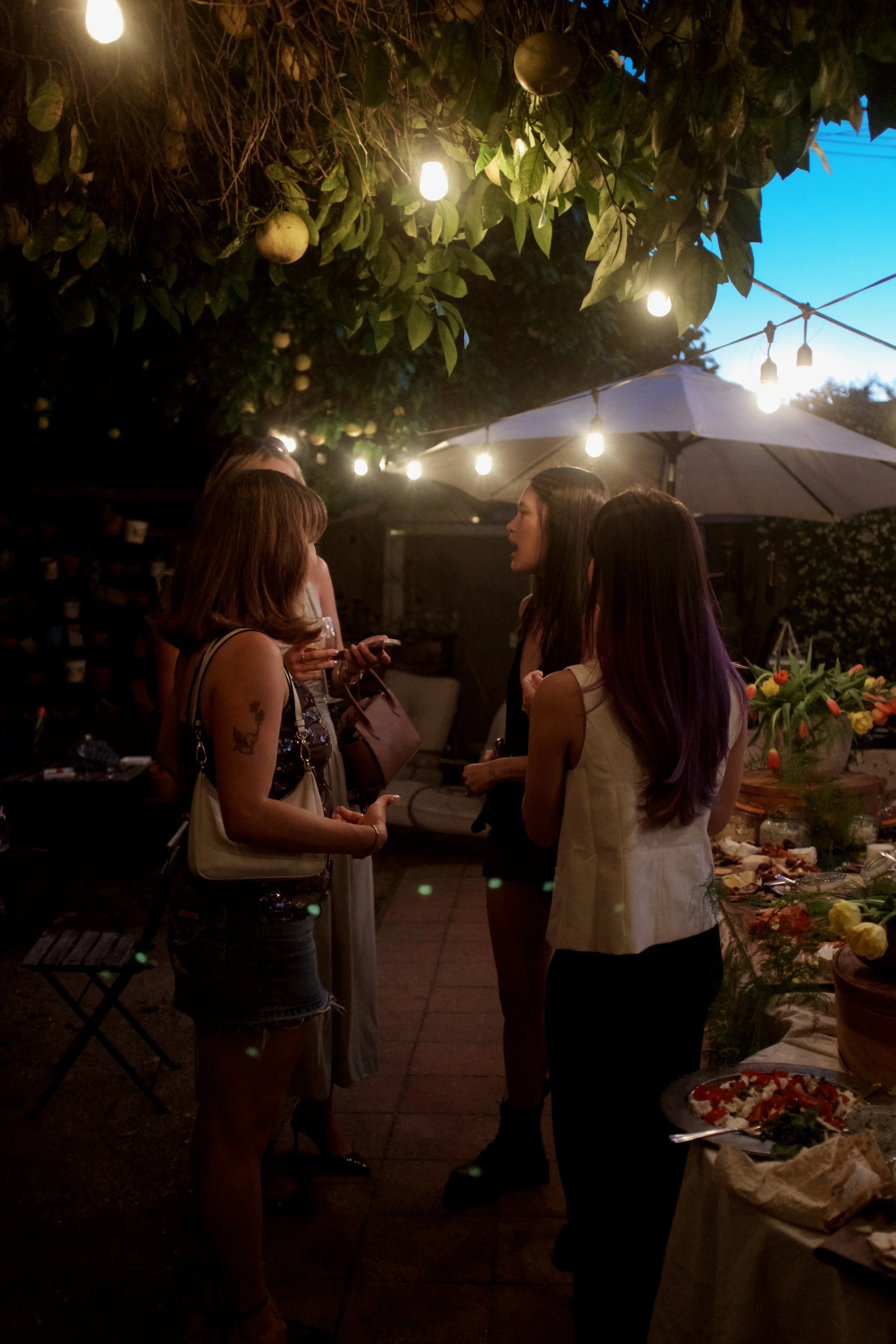 A group of women gathered at a nighttime outdoor party under string lights, with a table of food nearby.