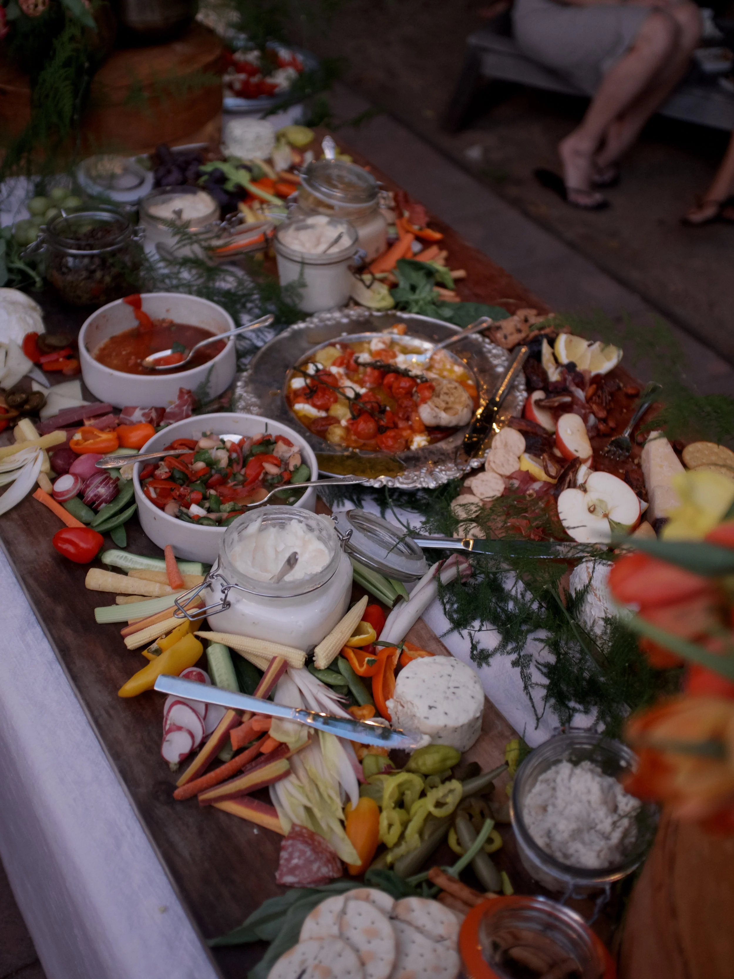A table of assorted foods including fresh vegetables, dips, and spreads, with some jars and bowls filled with various ingredients, at an outdoor gathering.