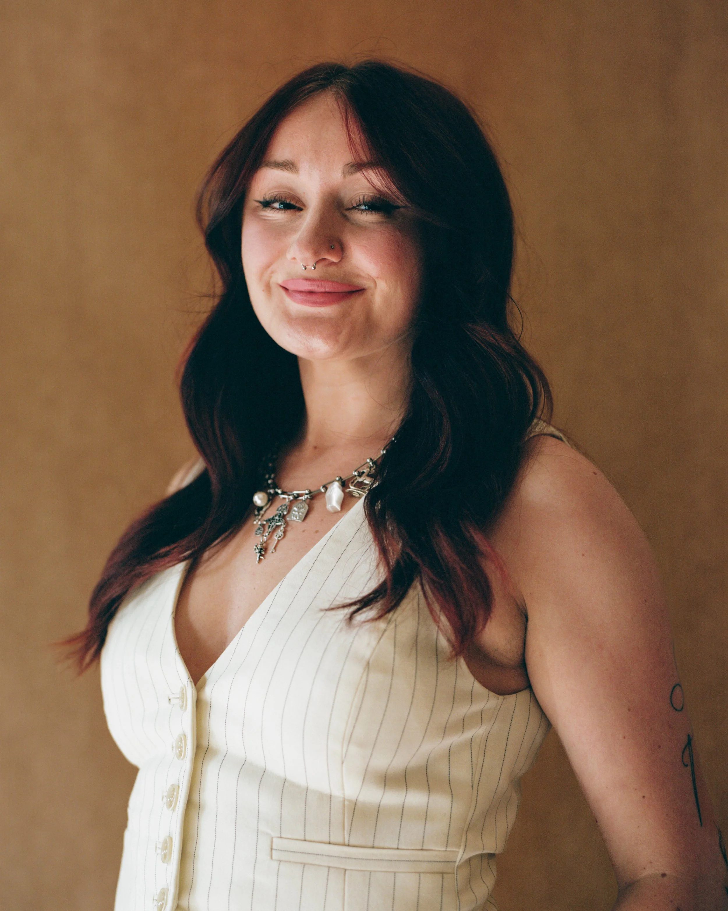 A woman with dark red hair, wavy, wearing a cream-colored sleeveless pinstripe top, a chunky silver and pearl necklace, and has a tattoo on her upper arm. She has a nose piercing and is smiling with one eye slightly closed against a plain brown background.