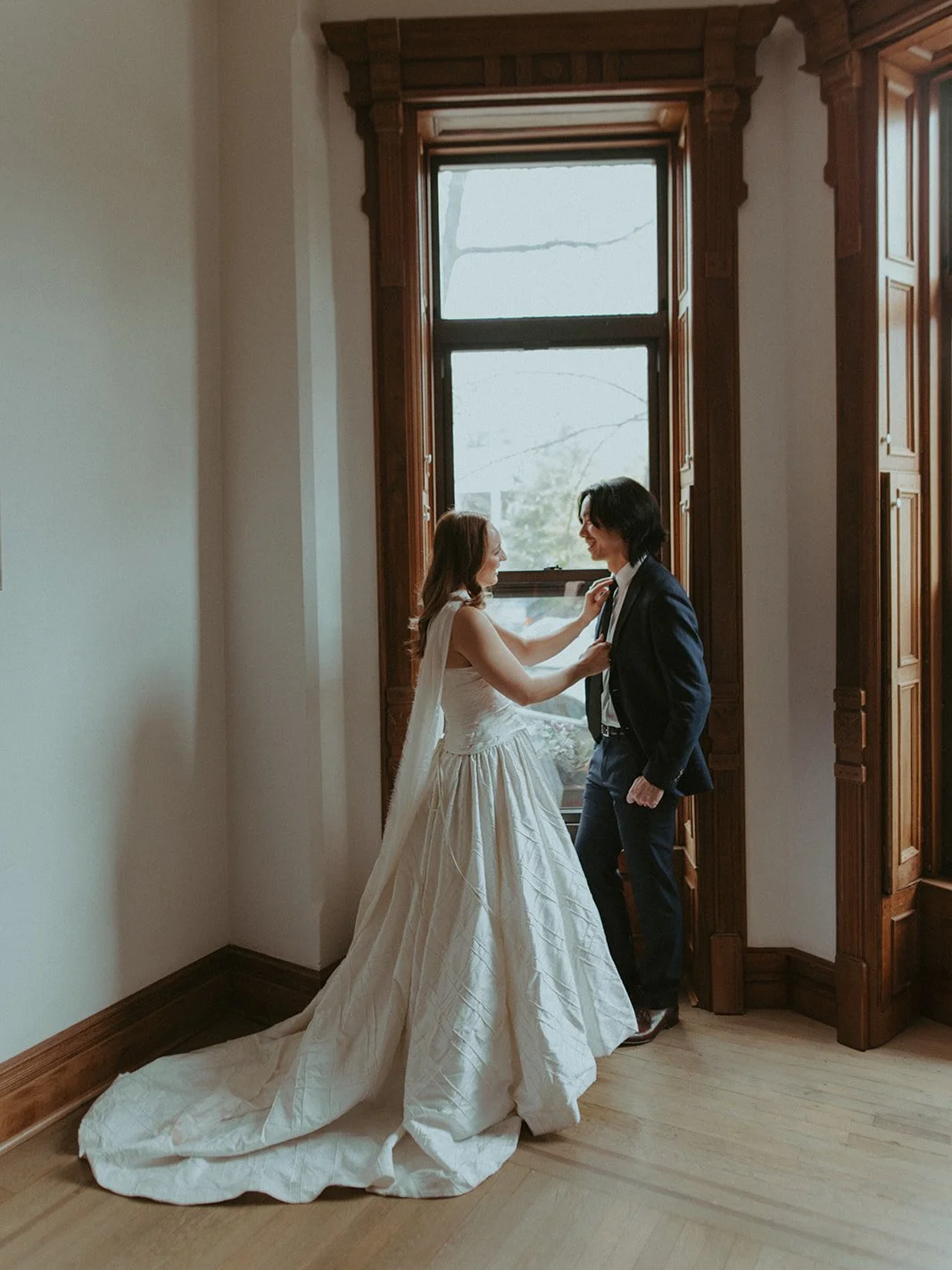 A bride and groom standing near a large window in a wooden-paneled room, smiling at each other.