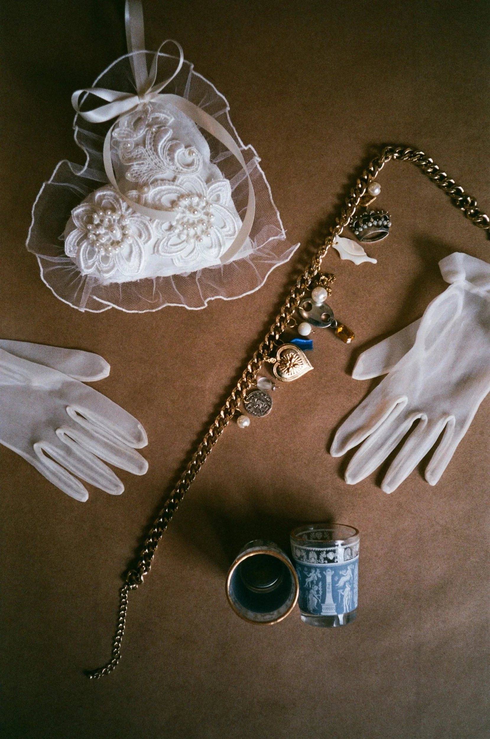 A collection of vintage jewelry and accessories, including a pearl bracelet, white gloves, a vintage cup, and a heart-shaped decorative item with fabric flowers and pearls, arranged on a brown surface.