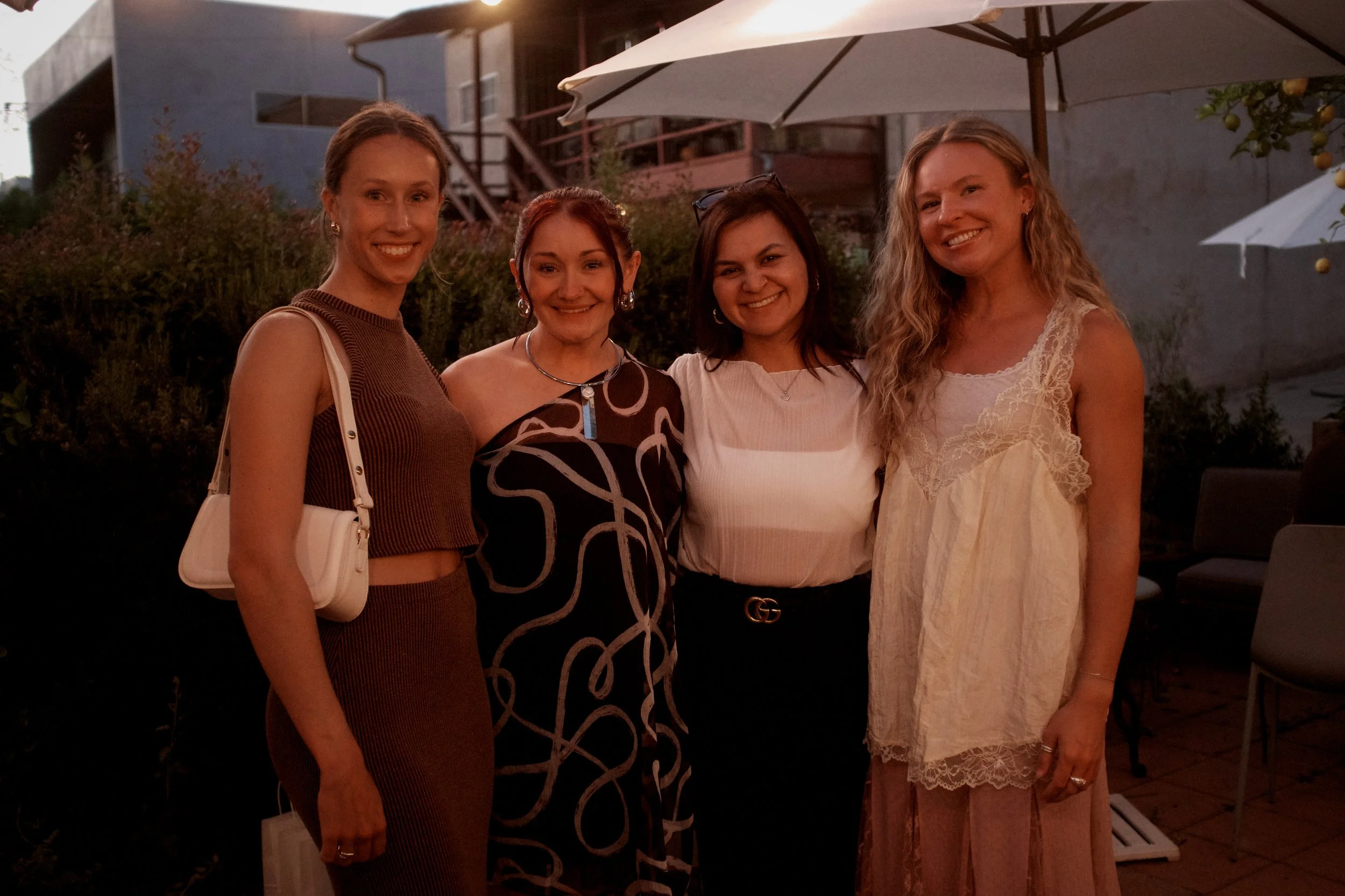 Four women standing together outdoors during evening time, smiling, with umbrellas and a building in the background.