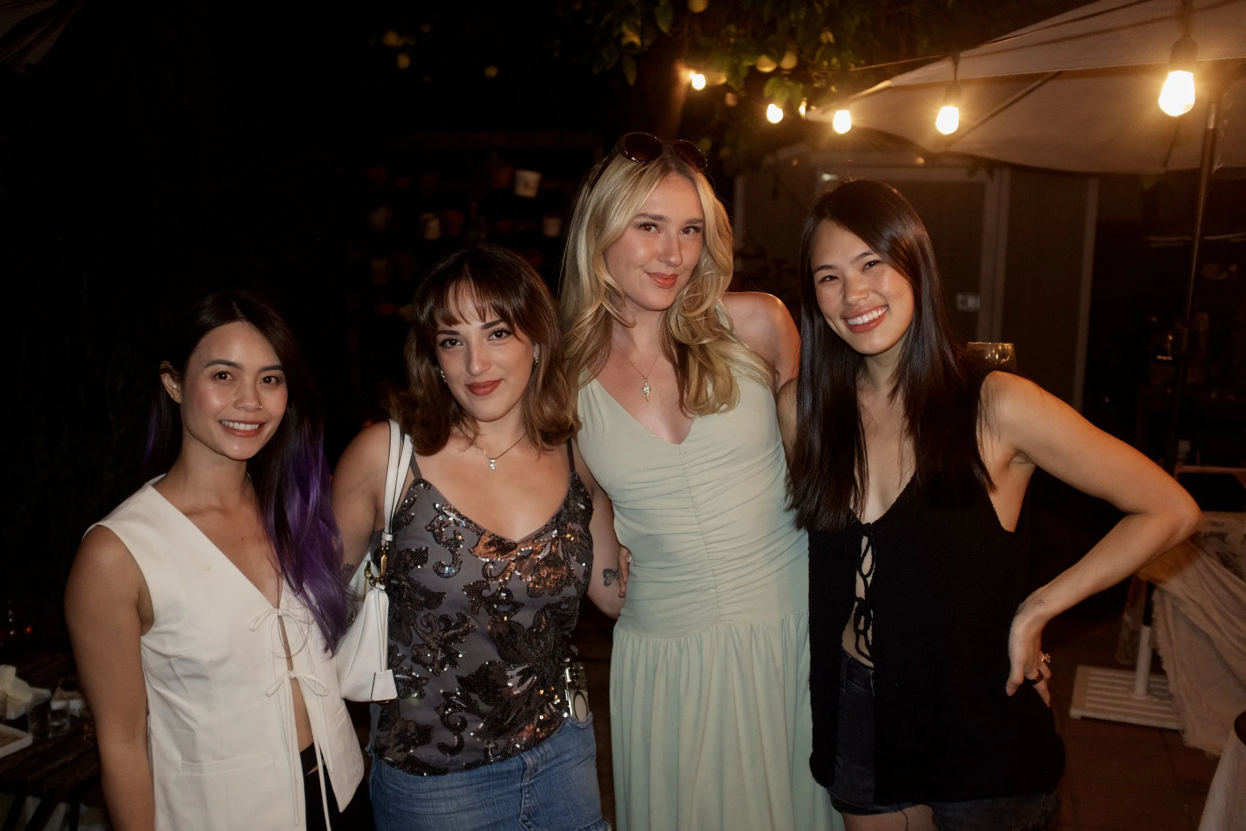 Four women standing together at night, smiling outdoors under string lights and a white umbrella.
