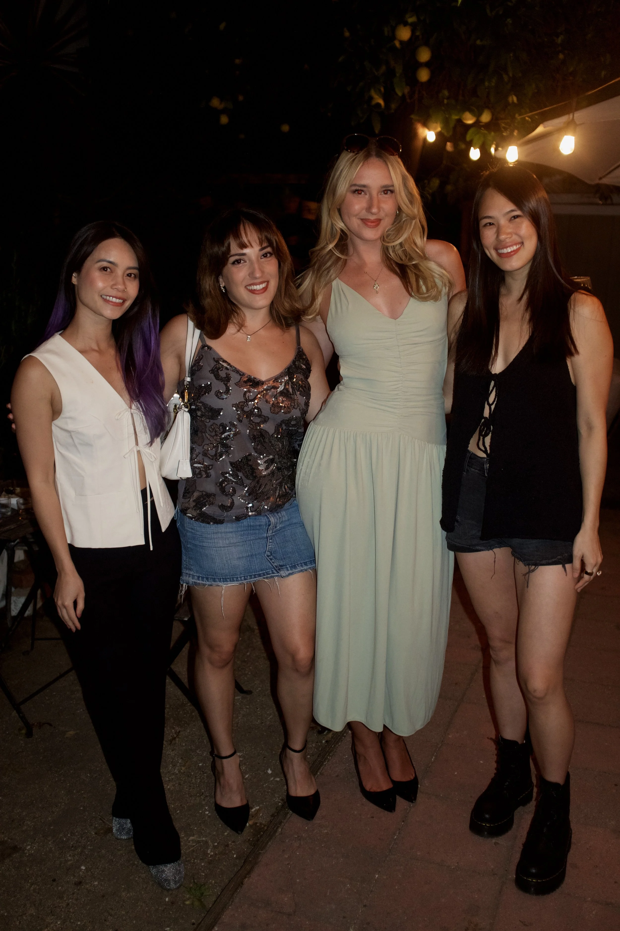 Group of five women standing together outside at night, smiling at the camera. The women are dressed in casual and dressy summer outfits. There are string lights and trees in the background.