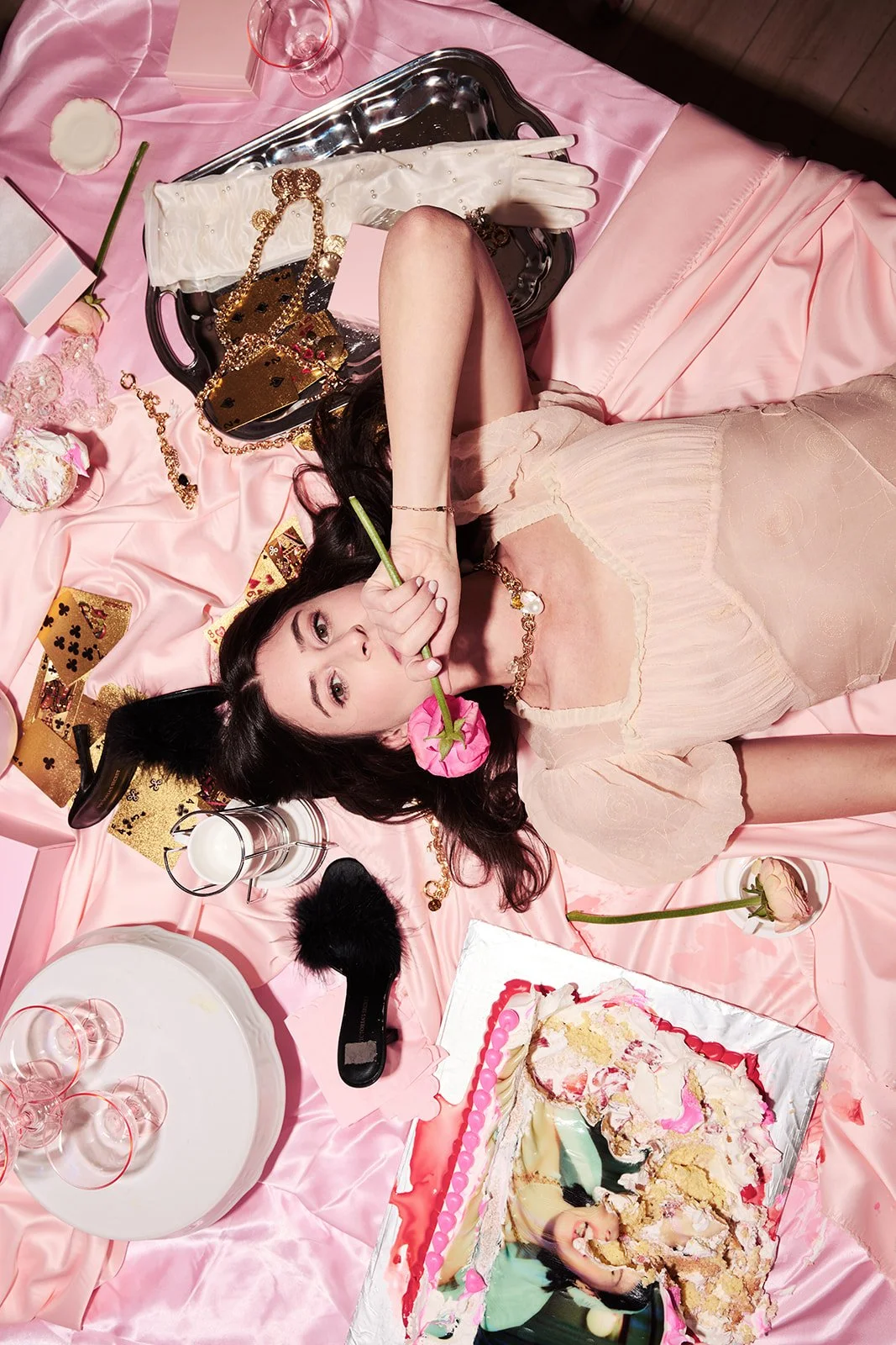 A woman lying on a pink satin sheet surrounded by party items, holding a pink rose in her mouth, with a partially eaten pink and white cake nearby.