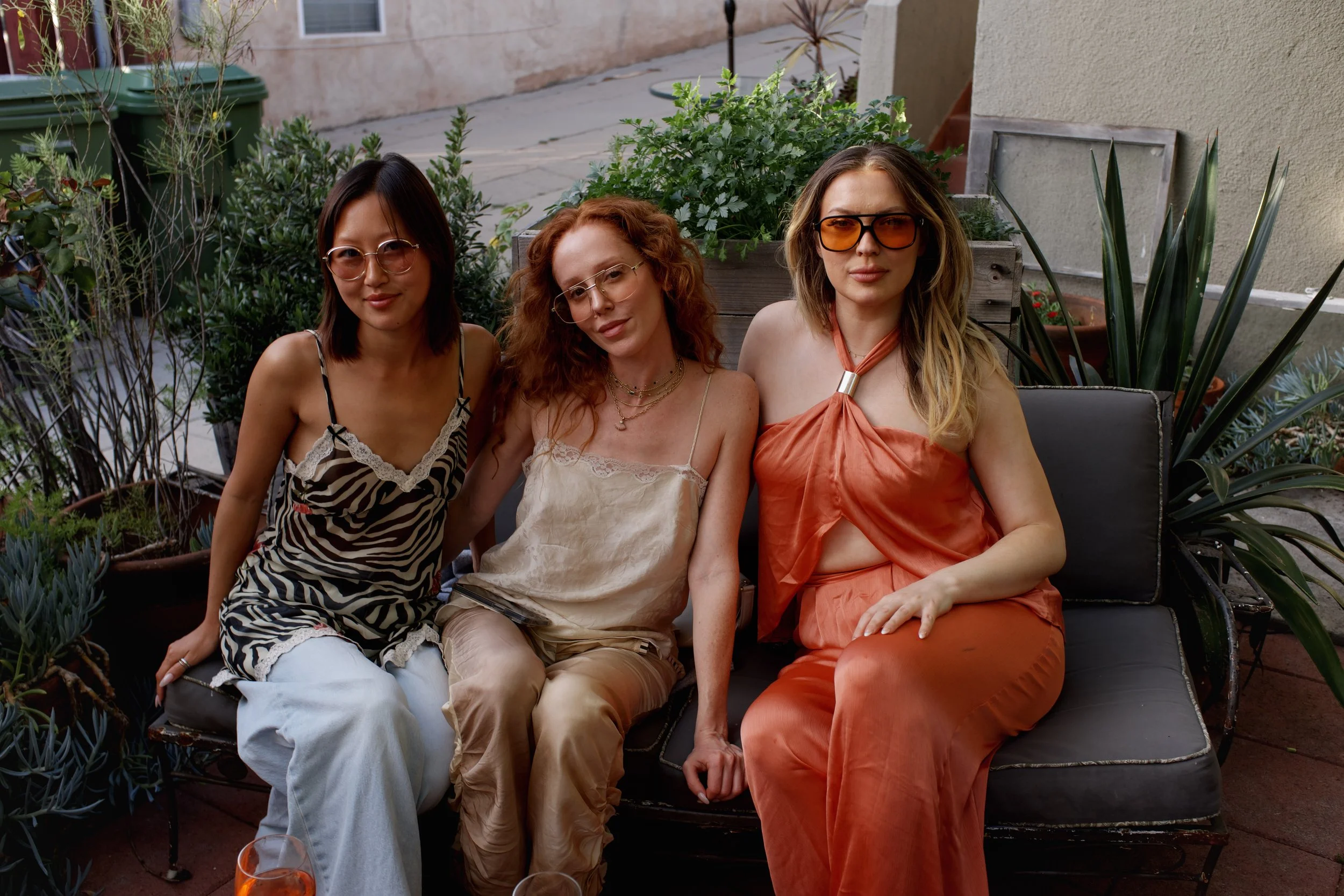 Three women sitting on outdoor sofa surrounded by green plants, all wearing fashionable sunglasses and casual summer clothing, smiling and relaxing.